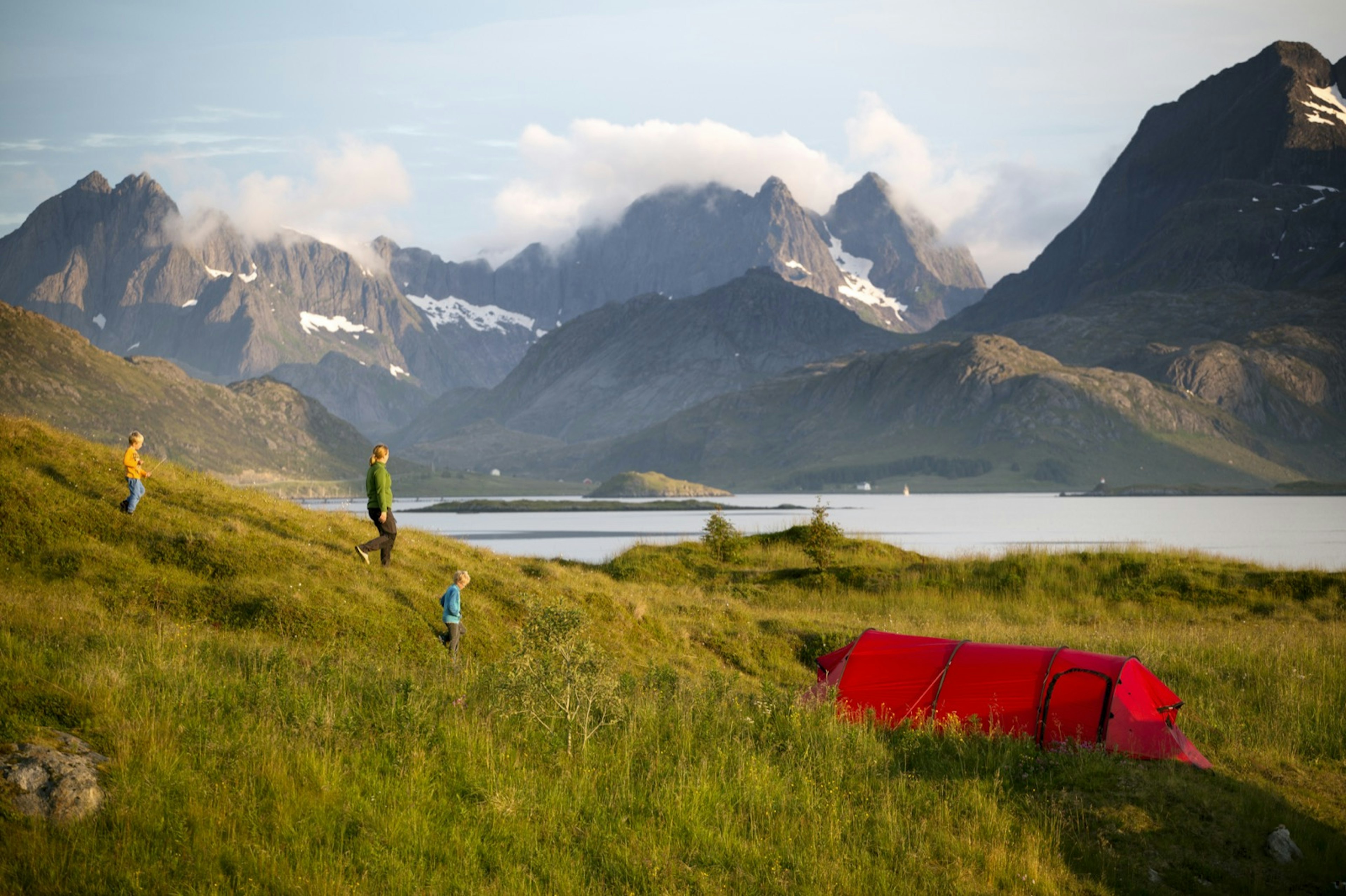 A mother and two children walk down a grassy hill toward a red tent with mountains and a body of water in the background