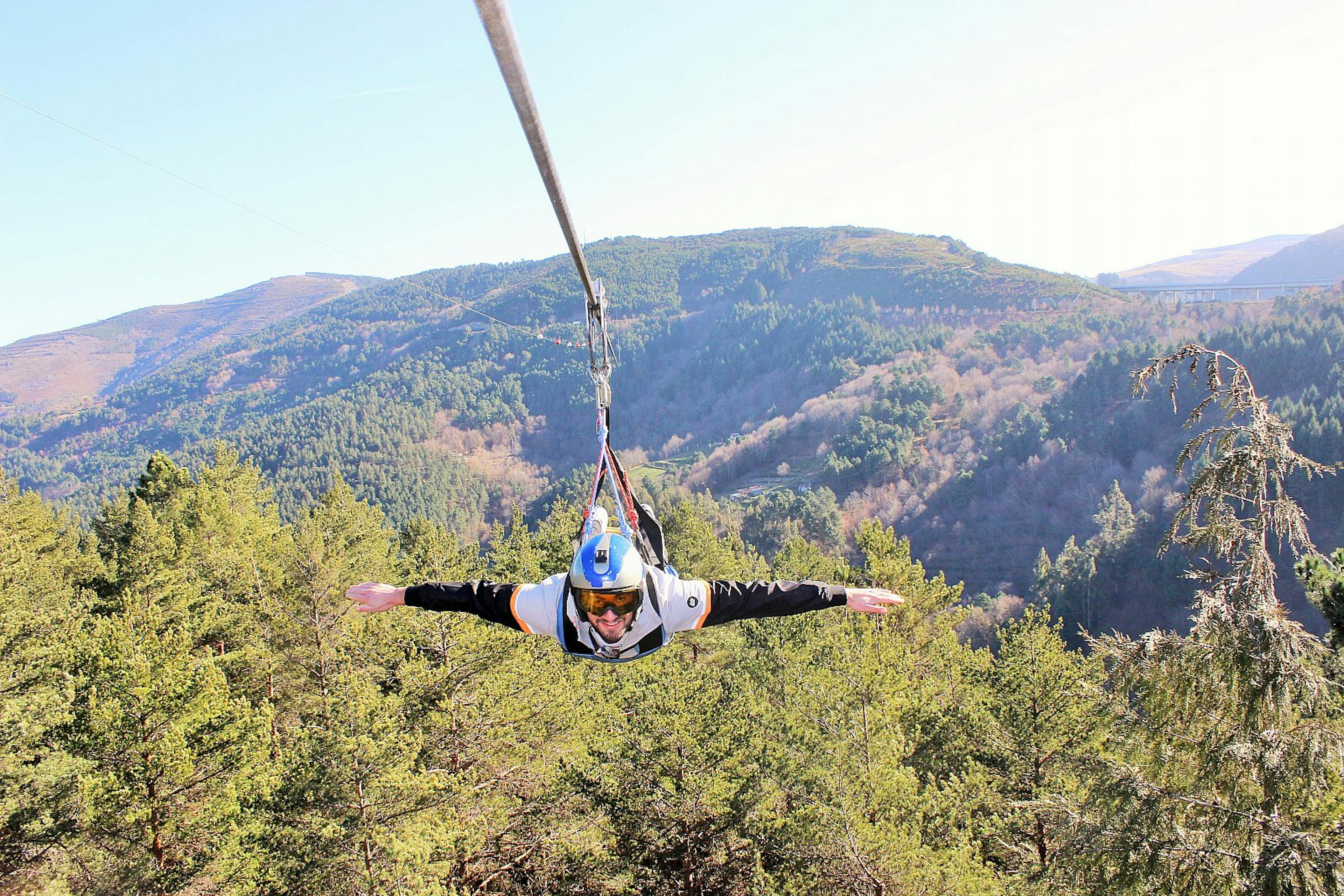 A man on a zip line at Pena Aventura, far above treetops with hills beyond; he is facing the camera with arms outstretched.