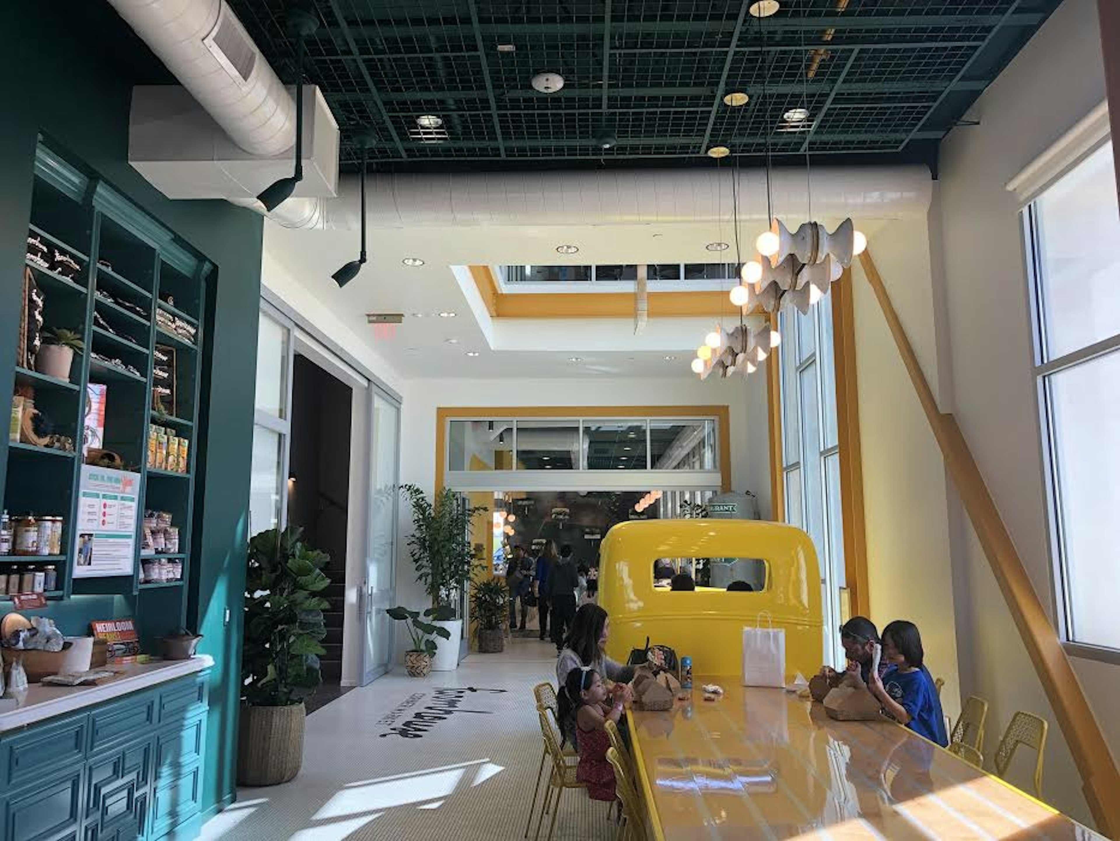 The interior of Farmhosue Corner Market includes a deep teal built-in cabinet on the left holding all kinds of tchotkes and kitchen items, and a long blonde wood table that ends in a yellow vintage truck cab. At the table, where the truck's bed once was, sits a family with small children. The white tile floor extends far back into a space full of plants and light with white walls.