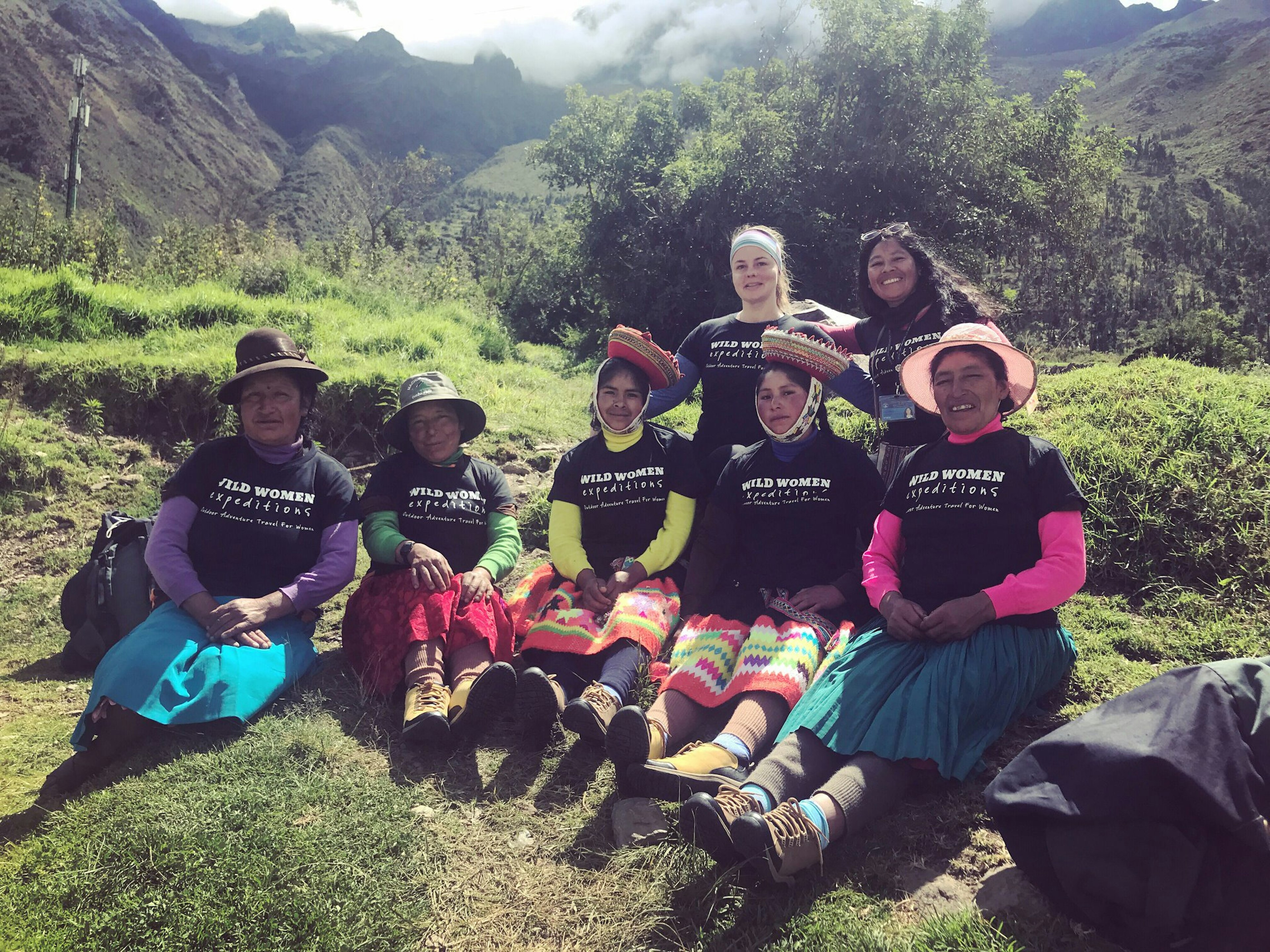 A group of female porters in Peru pose for the camera on a grassy hill along the Inca Trail. They are wearing black shirts that say 'Wild Women Expeditions' over their long skirts and long-sleeve shirts; women adventure travel