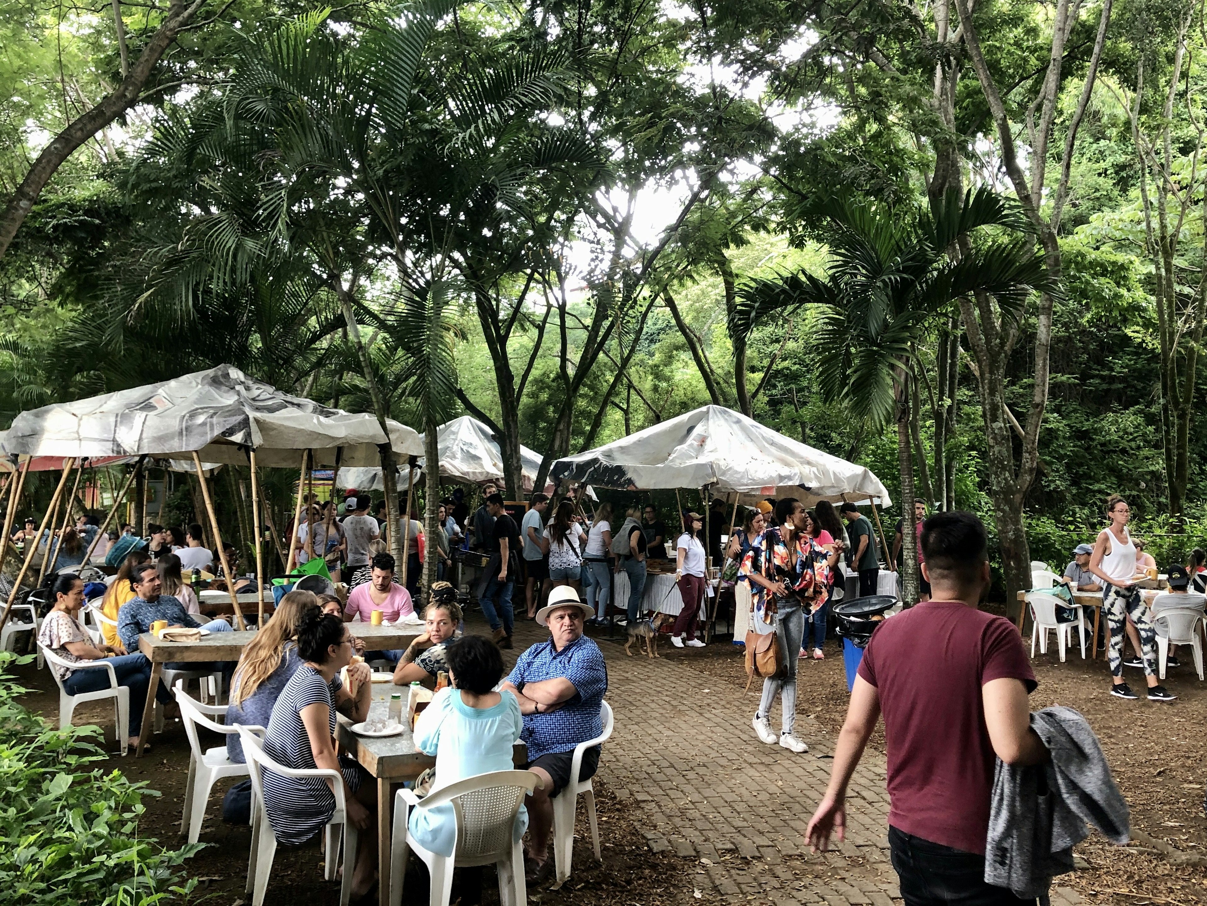 Large groups of people sit at a collection of tables while others stand by outdoor stalls on a paved brick lane. There are tall, leafy trees framing the photo.