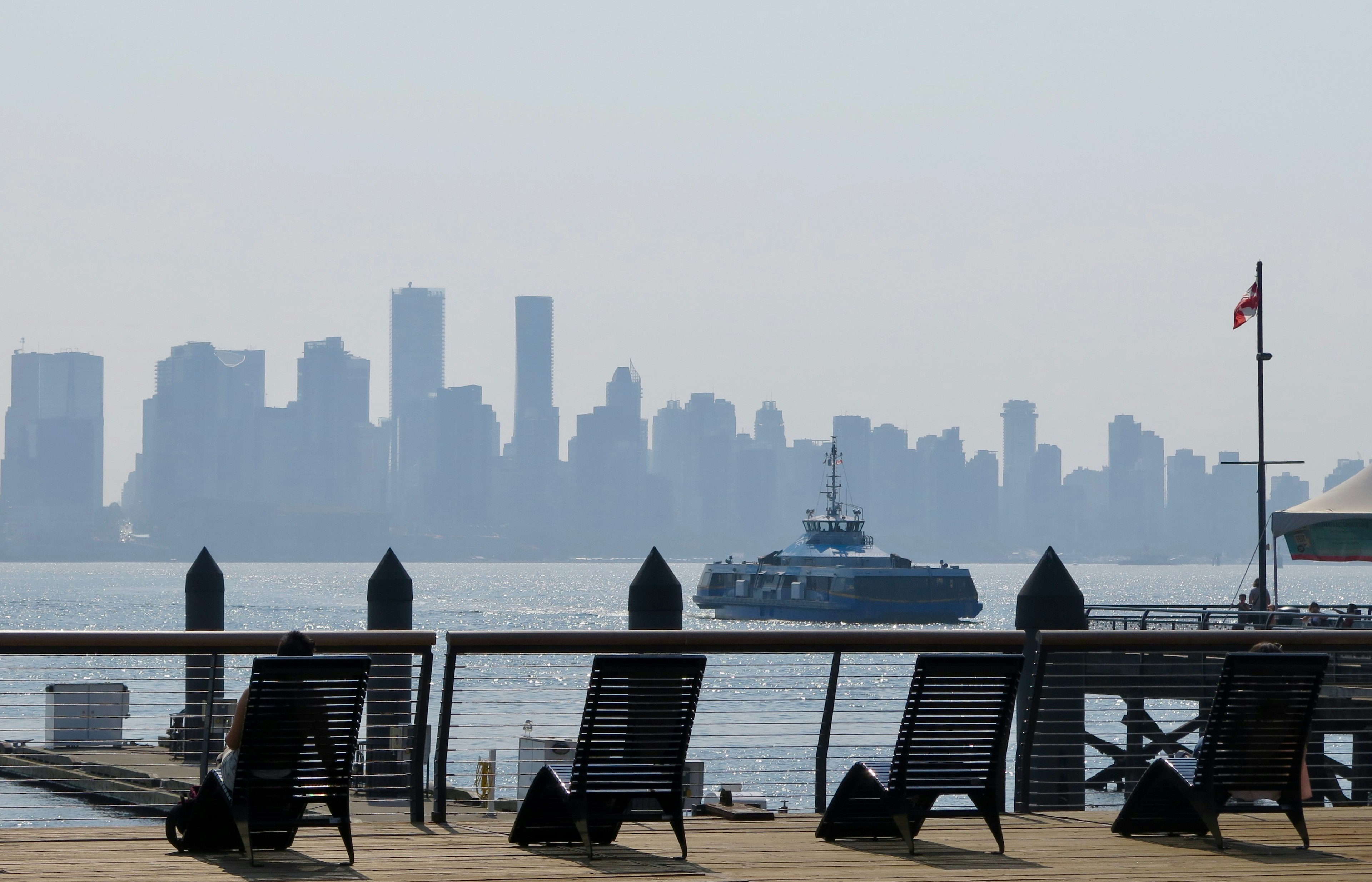 A woman sits in a reclining lounge chair next to a railing overlooking a waterway in Vancouver. A ferry sails through the waterway toward the far shore which is dominated by skyscrapers.