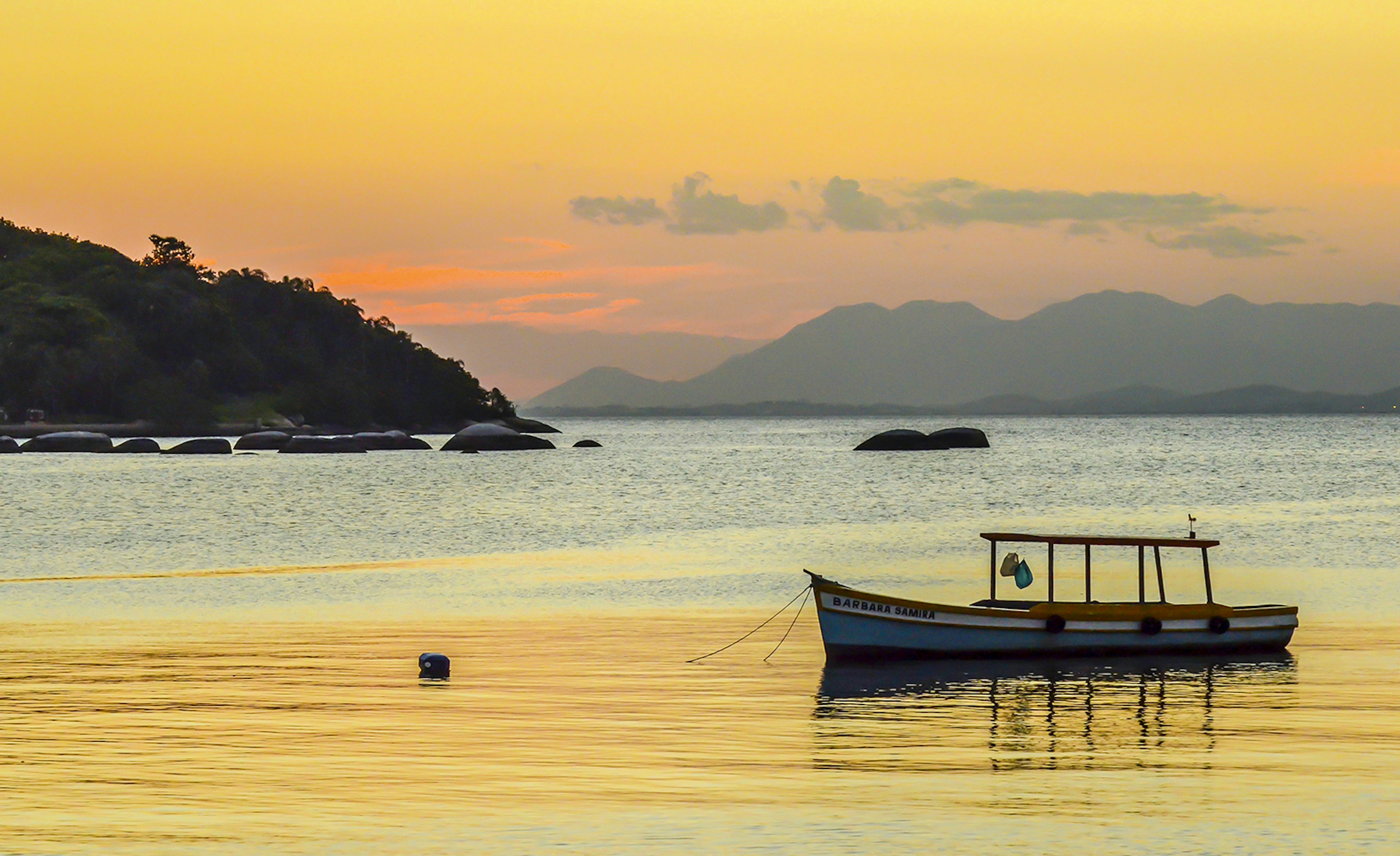 A small fishing boat is moored in glassy water that reflects the orange sky during sunset. Ilha da ʲܱá, Brazil.