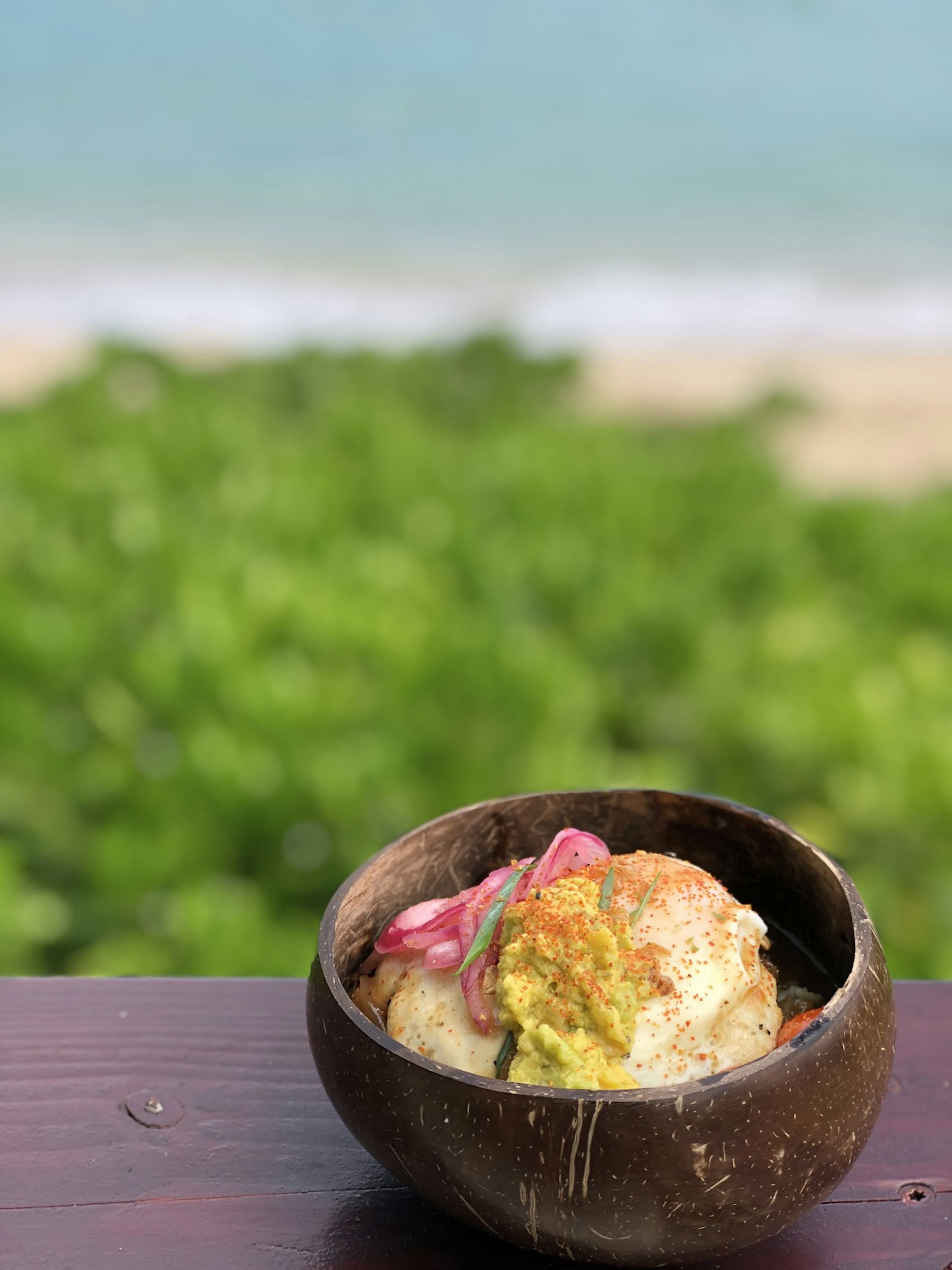 A small brown bowl - possibly a coconut - has some hawaiian food in it. You can see a beach in the background