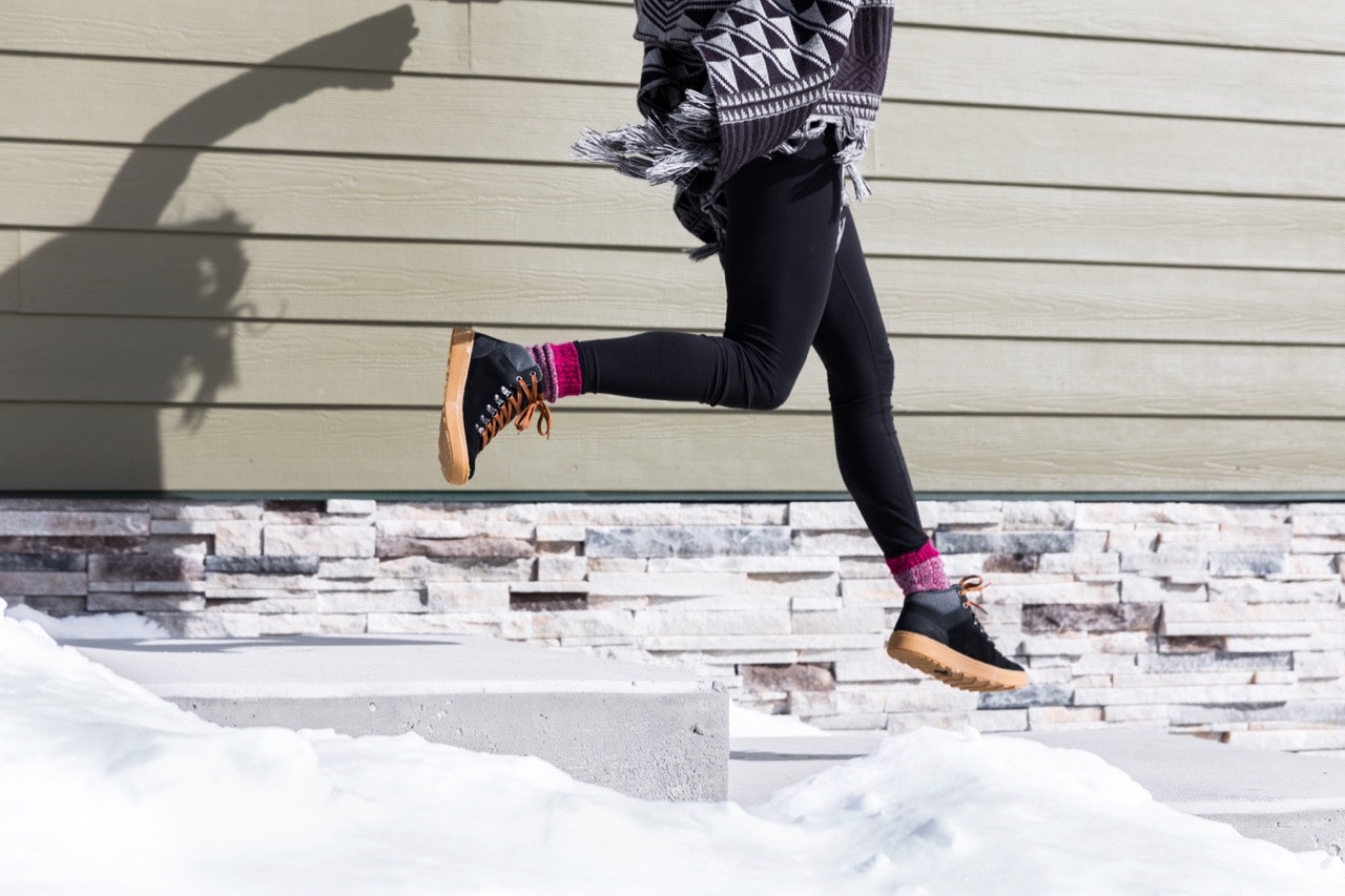 a woman jumping down snow-covered steps wearing Forsake's Lucie Mid boot in black