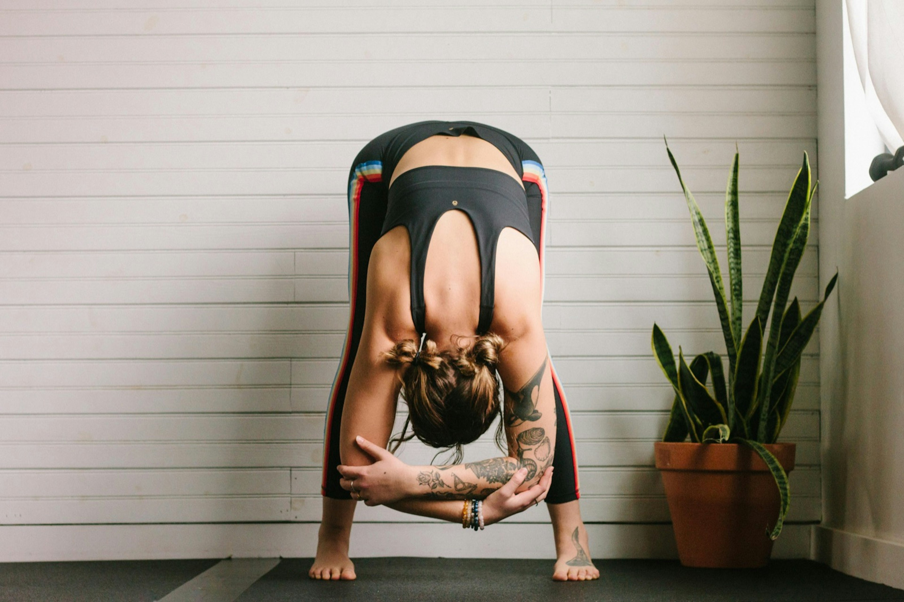 a woman in black performs a forward fold yoga pose on the backdrop of a white wall