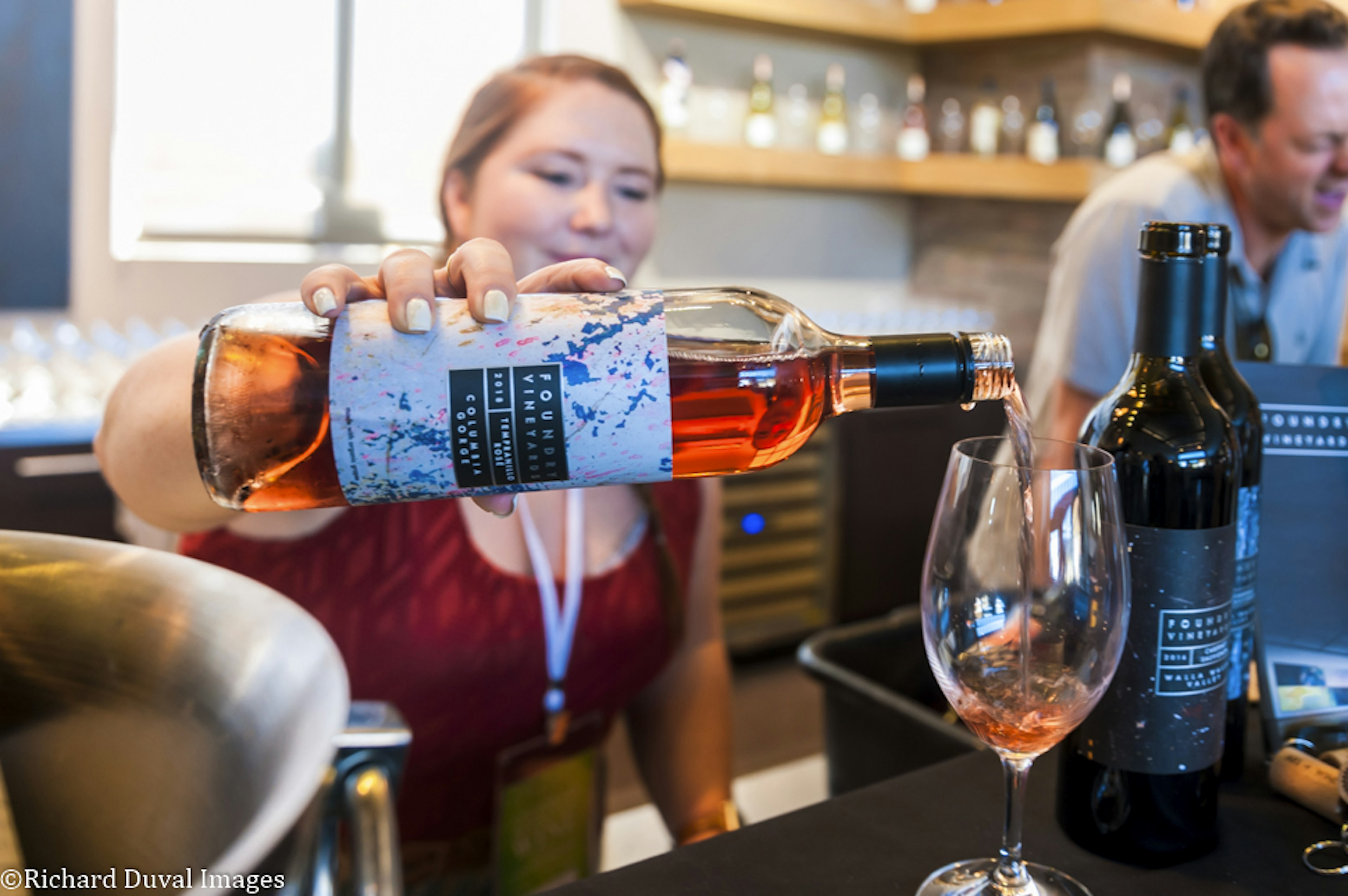 A woman pours a glass of rose-colored wine. Next to the wine glass are dark-colored bottles of Foundry wine and a man talking; Walla Walla wine