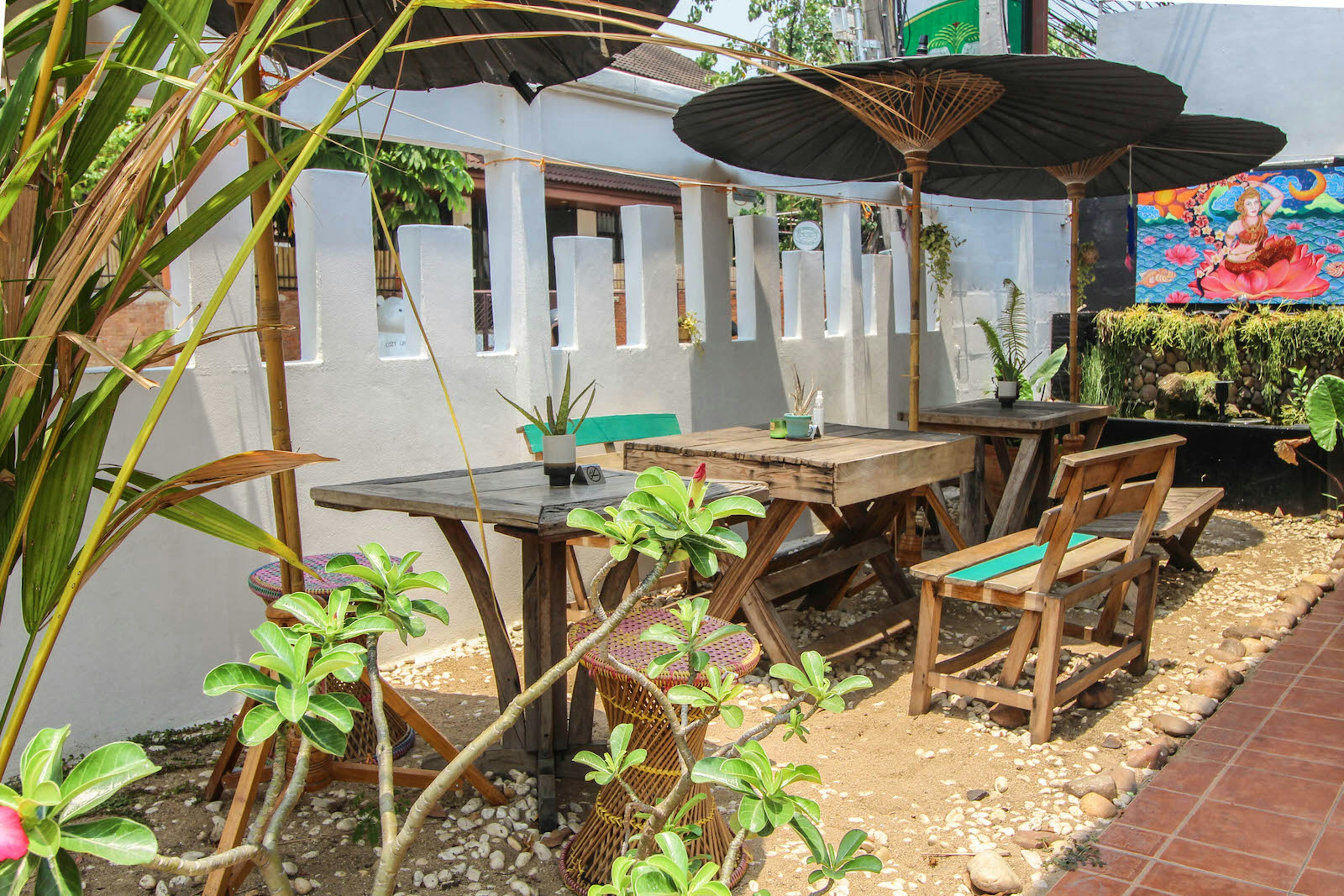 The outside seating area at Free Bird Cafe in Chiang Mai. Wooden tables and chairs are shaded by straw parasols. There is a colourful mural on the far wall.