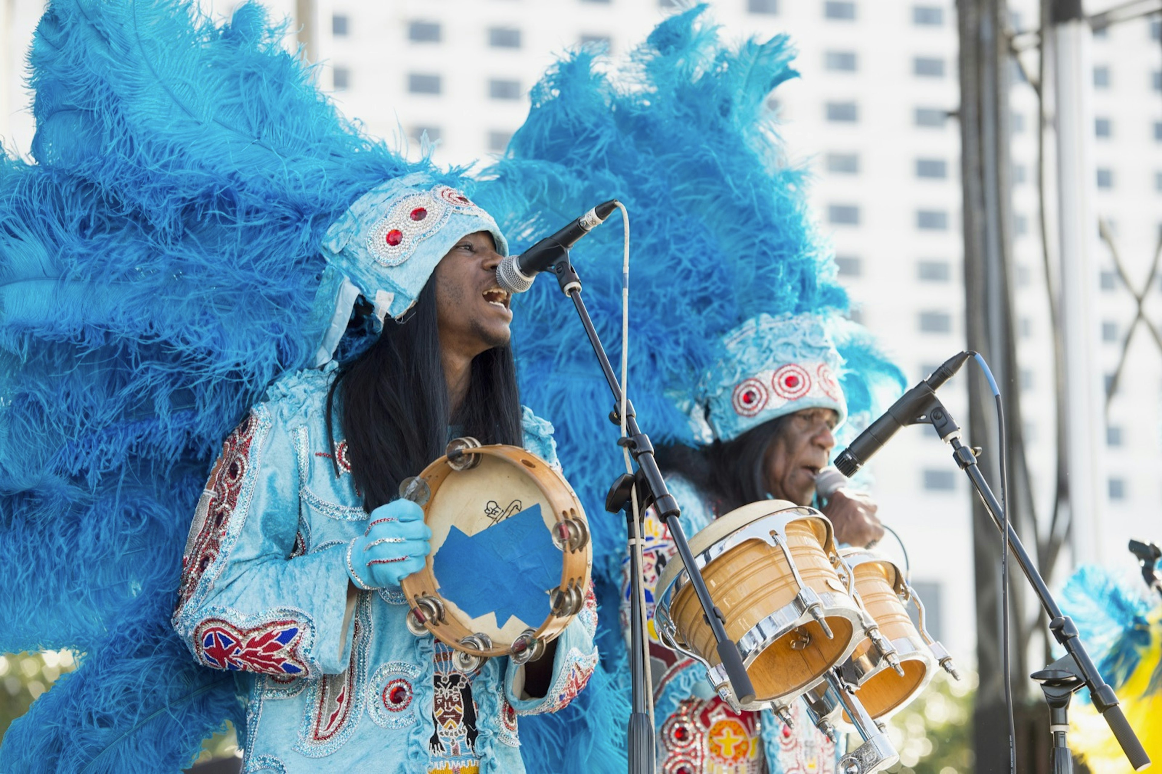 Two Mardi Gras indians in light blue feathered costumes play a tambourine and bongos on a stage; New Orleans Festivals