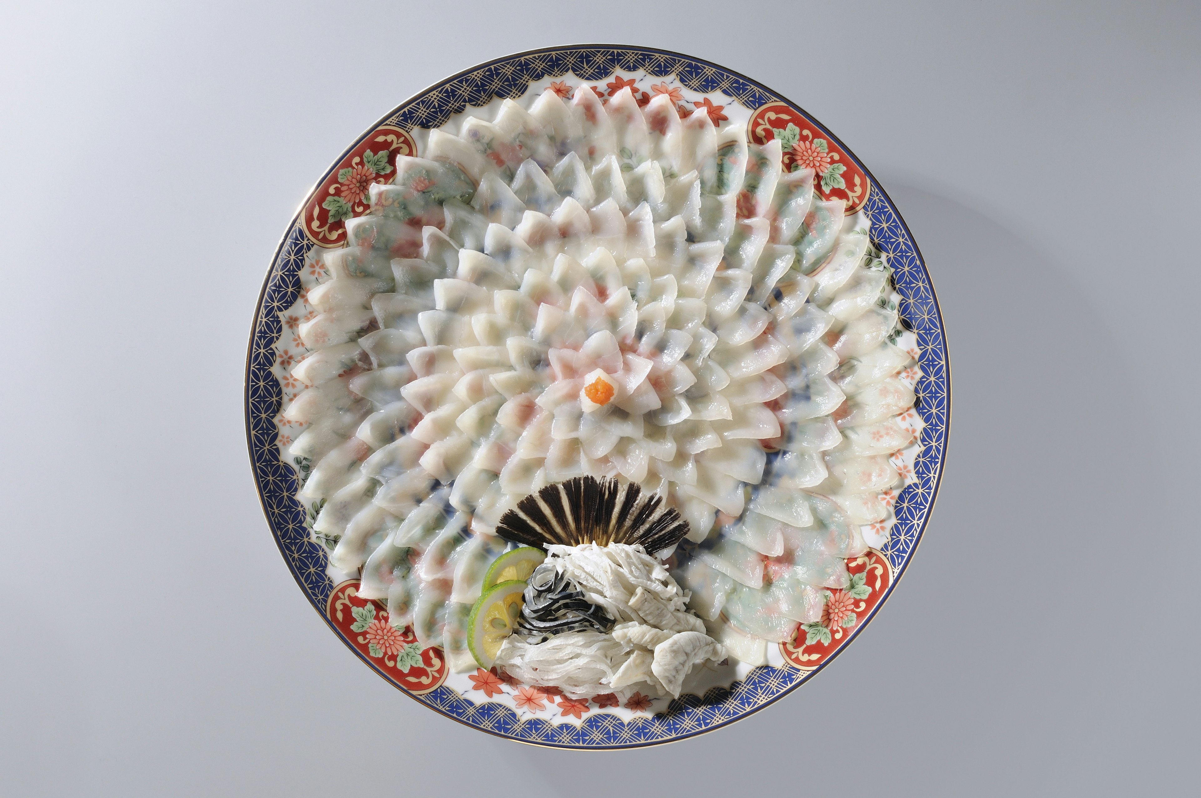 Thinly sliced fugu (puffer fish) is intricately arranged in the shape of a flower on a colorful plate.