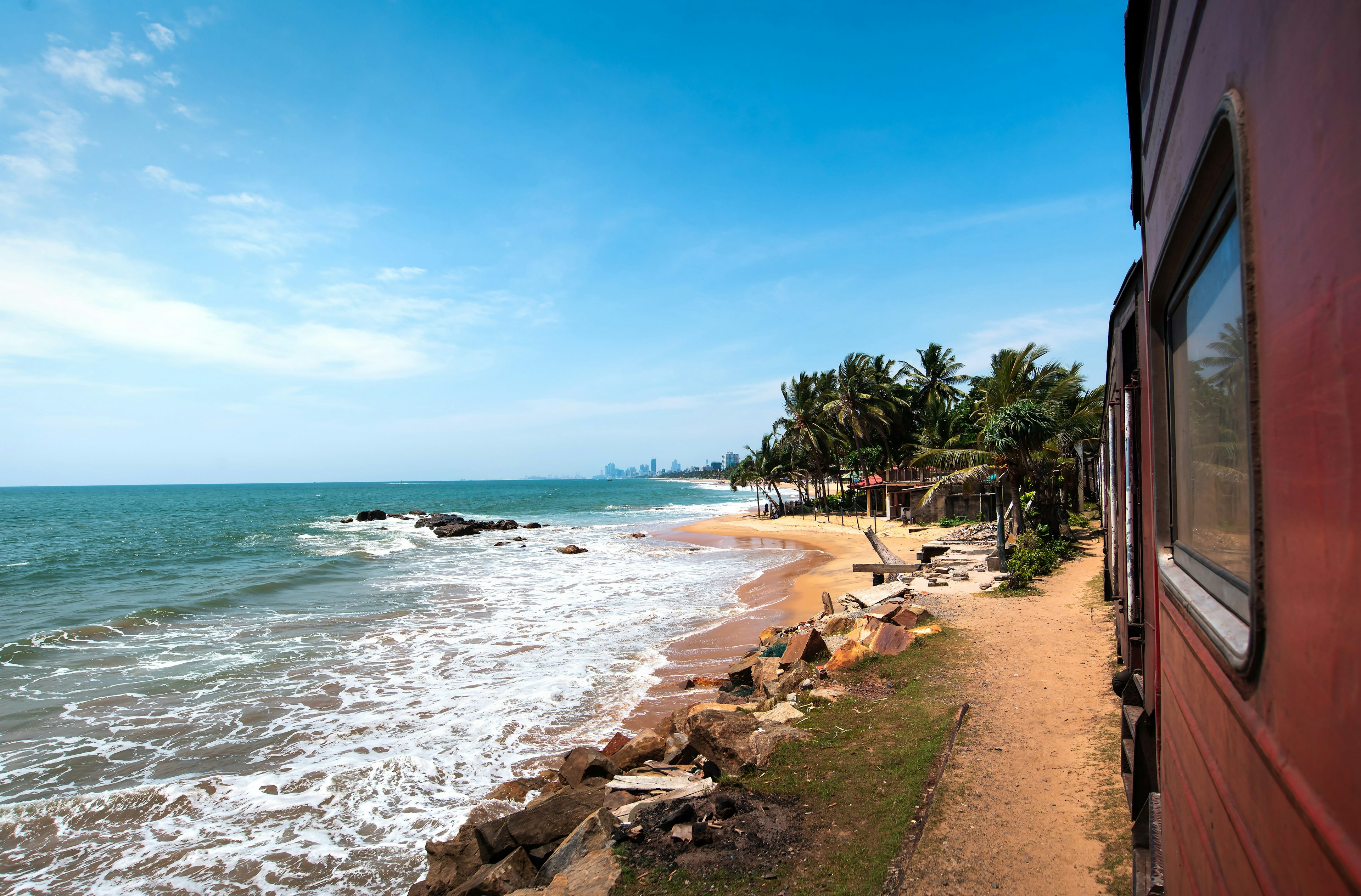 A parallel view of a red train travelling from Colombo to Galle, taken by a passenger on board the train. The tracks runs alongside the coast, with the sea coming right up to the tracks. A small, palm tree-backed beach is visible in the middle ground, while in the distance, the skyscrapers of a metropolis, presumably Colombo, can be seen.