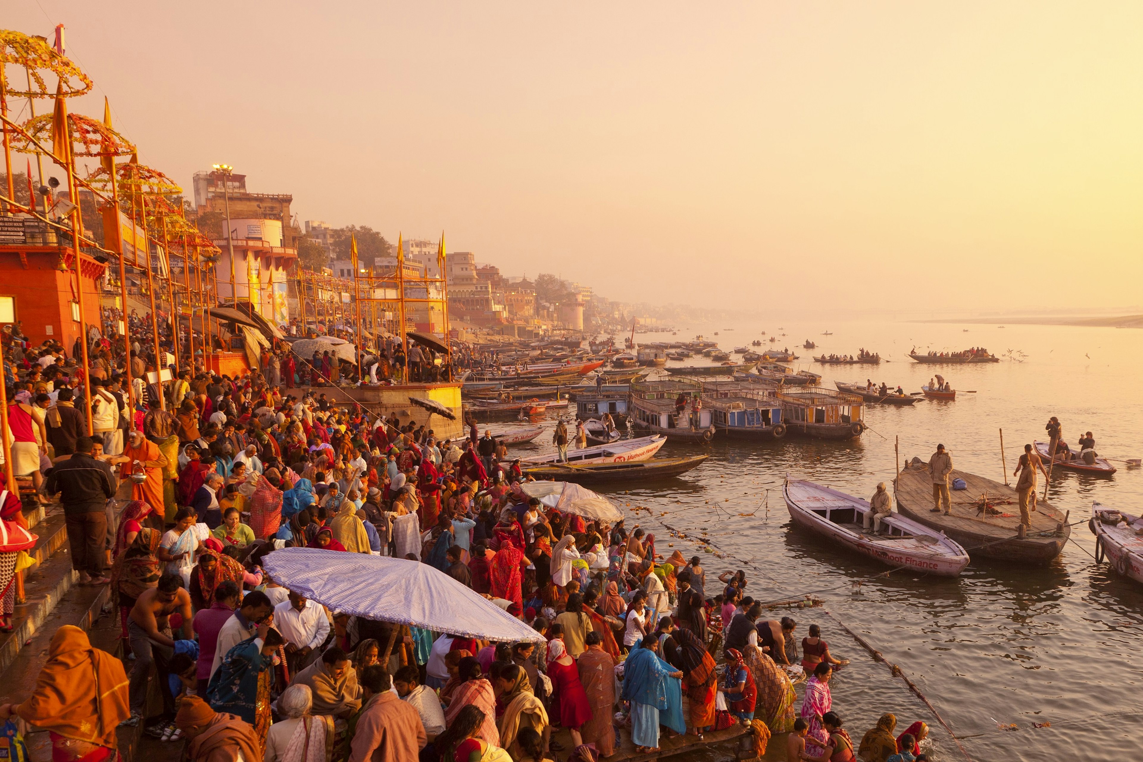 A shot looking down the River Ganges from the upper ghats; the steps down to the river are heaving with crowds for as far as the eye can see. Everything is rich in yellows, oranges and reds.