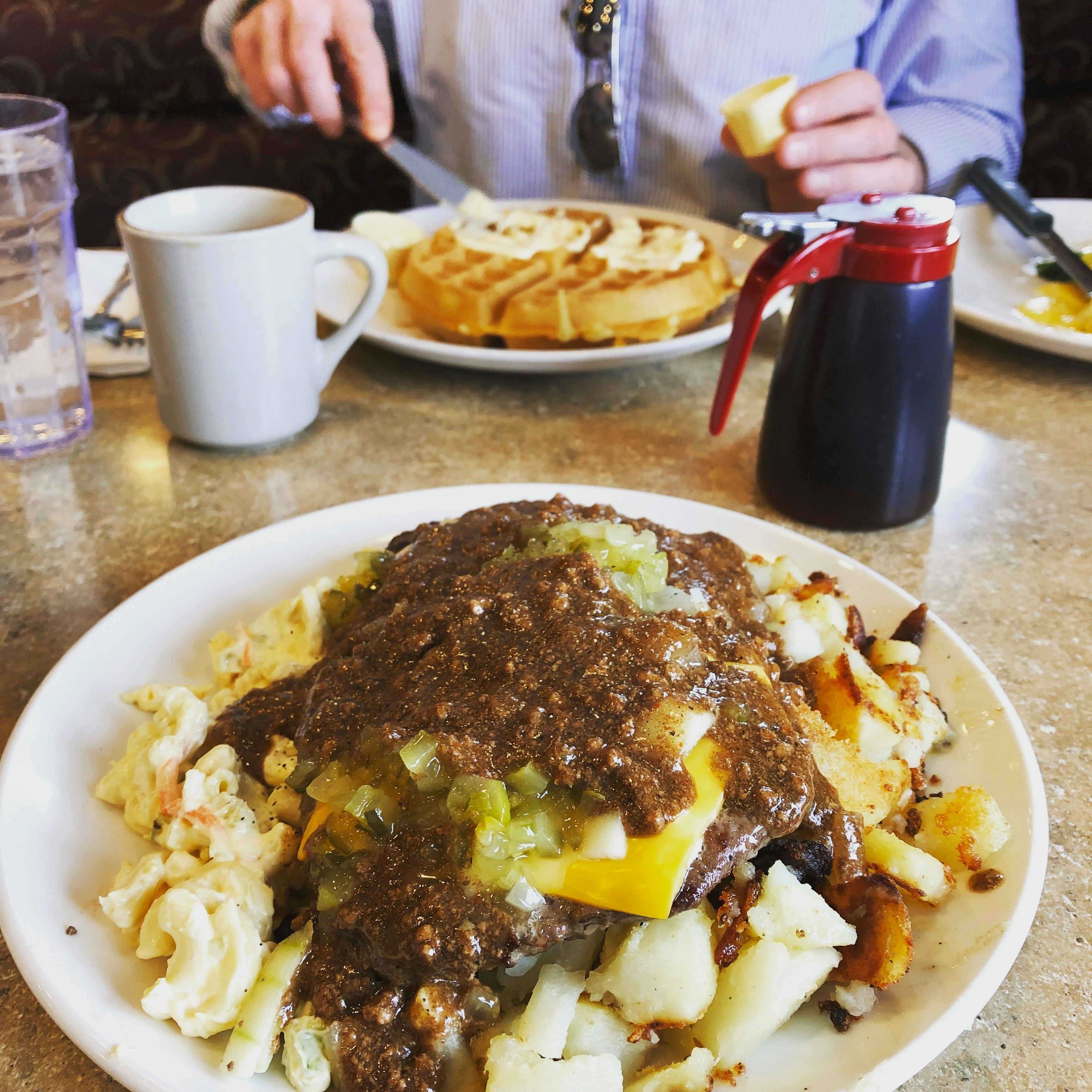 Closeup of a plate filled with macaroni salad, potatoes, cheese, gravy and bunch of other unidentifiable food items. Across you can see a person buttering a waffle. There is a small container of syrup in front of the plate.