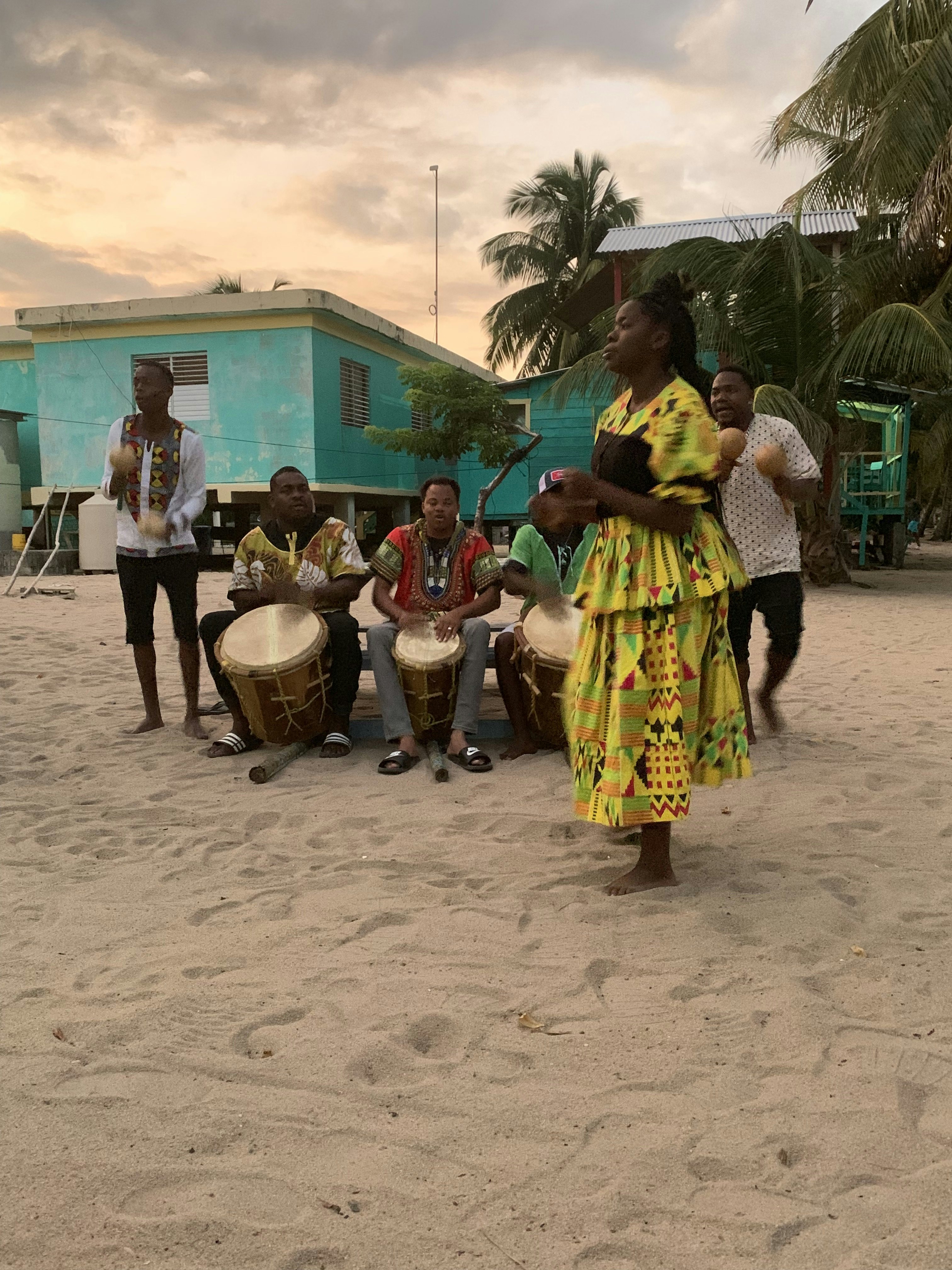 A trio of drummers are flanked by a pair of men playing a pair of maracas. There is a woman dancing in front of the musicians.