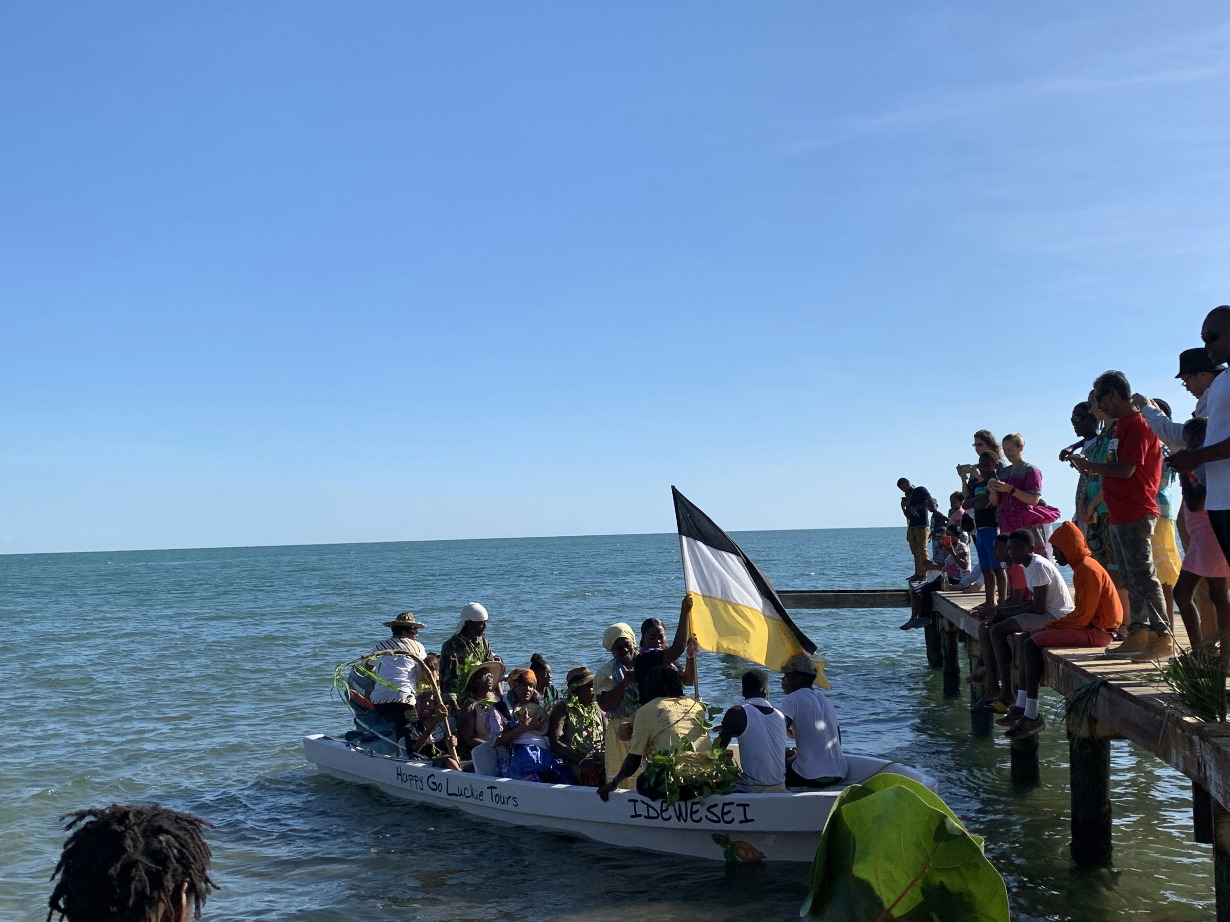 A large boat filled with actors re-enact Garifuna Settlement Day as a group of people watch from a wooden pier.