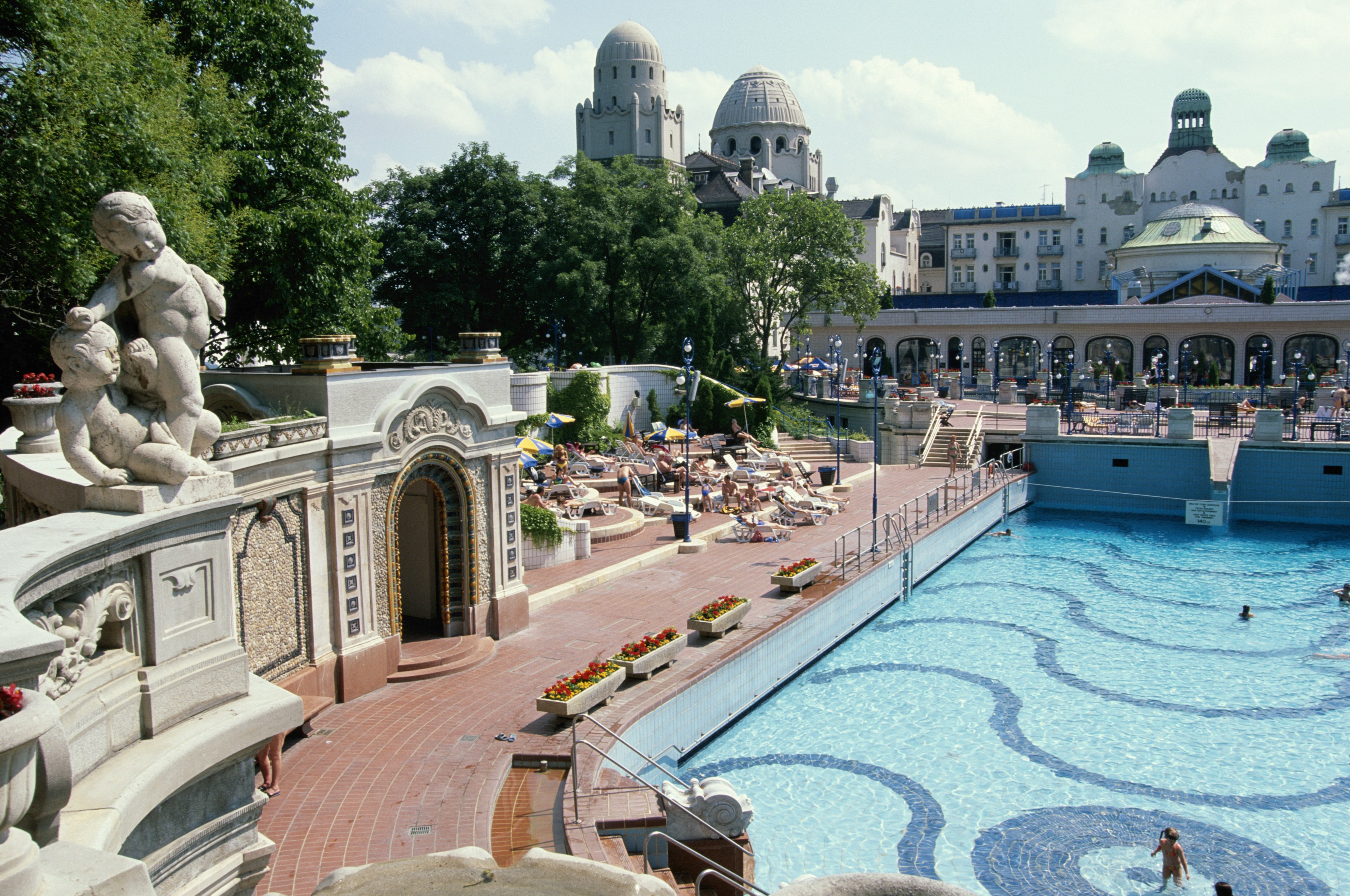 Stone pillars line a thermal pool; there are metal terraces with plants on them above the pool and a glass roof allowing for sun rays to peek through; Budapest autumn