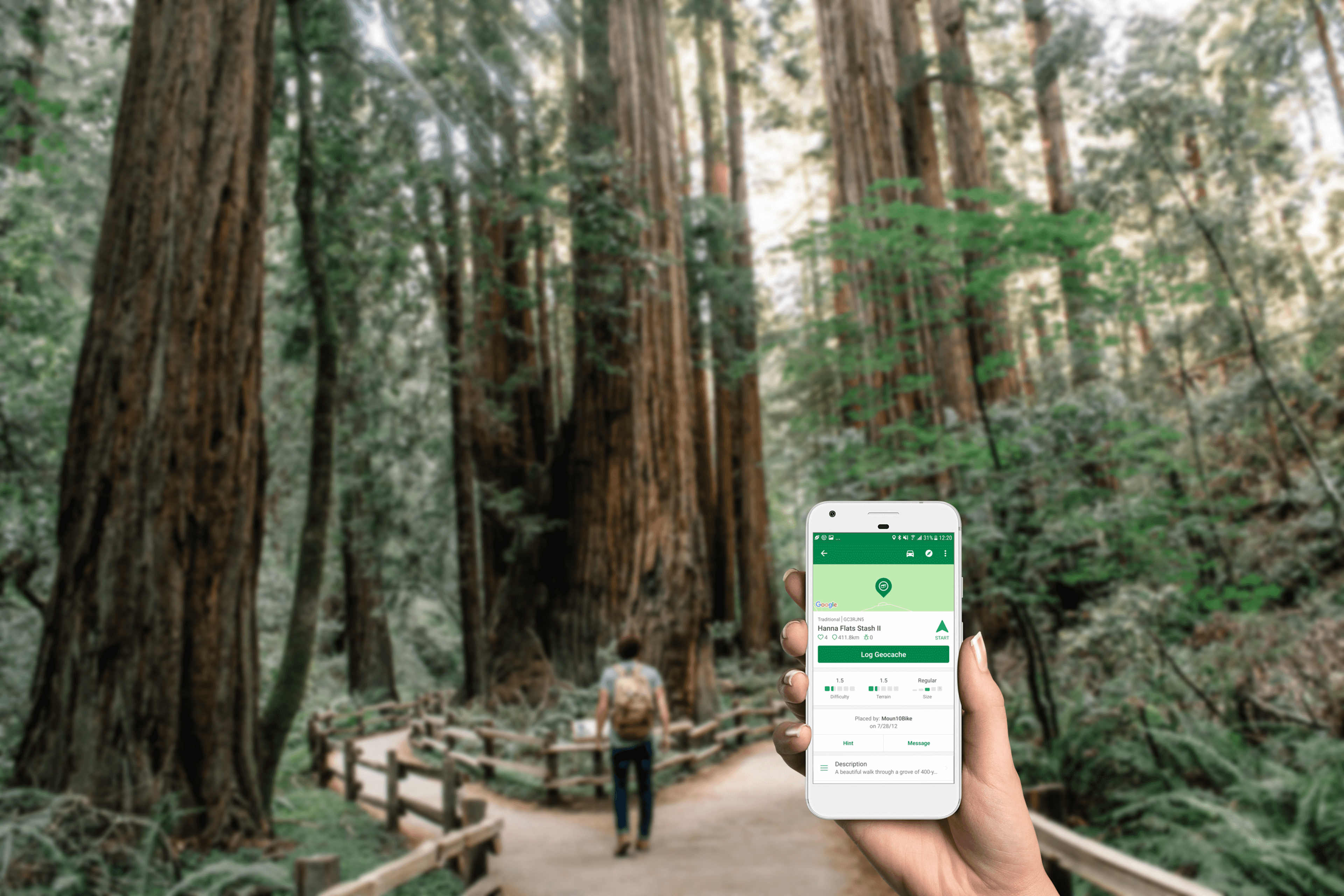 A hand holds up a smartphone, displaying the Geocaching App. A wooded area is visible in the background, with pathways and wooden fencing.