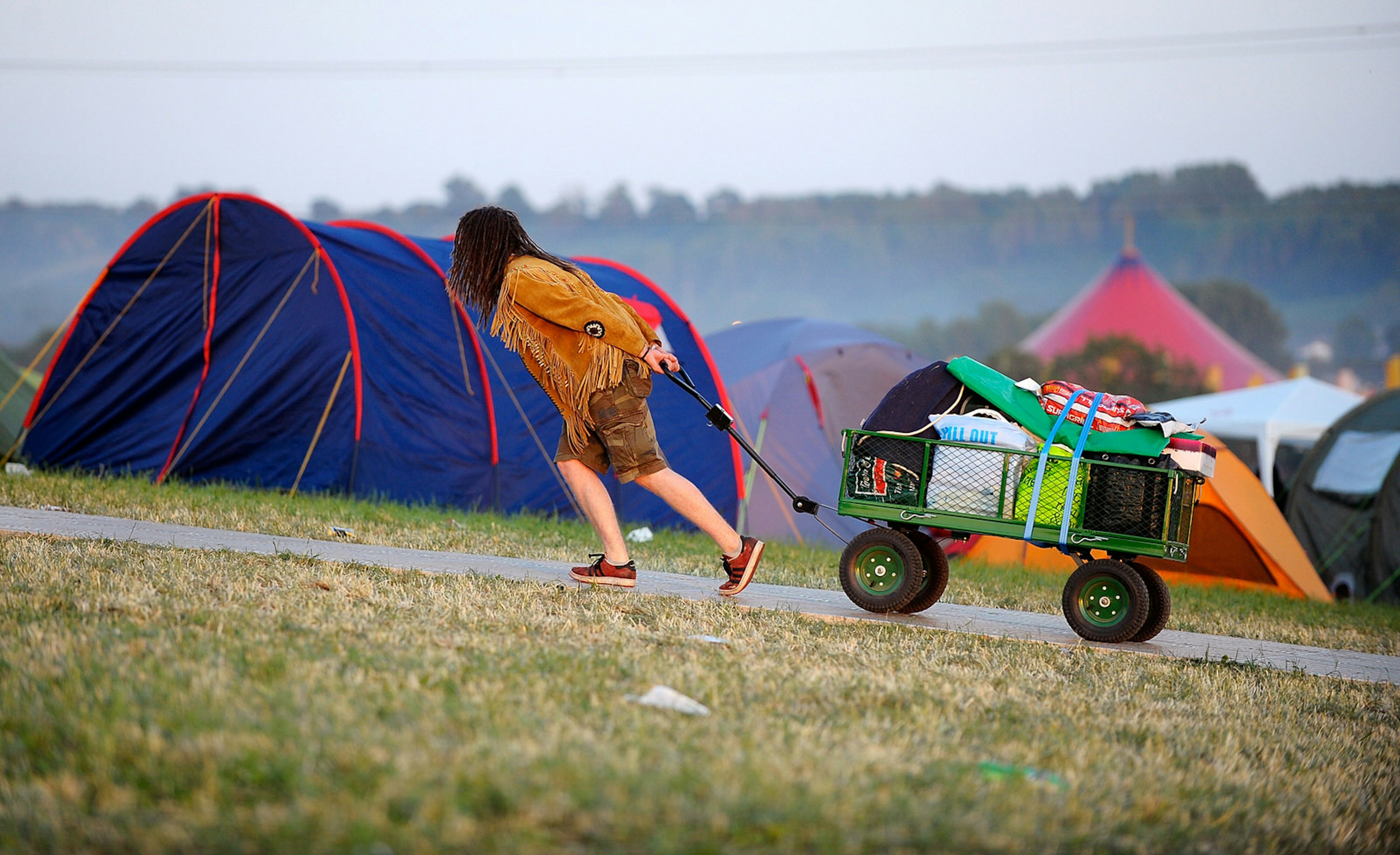 A festival goer drags a fully loaded trolley full of camping stuff up a hill at a campsite