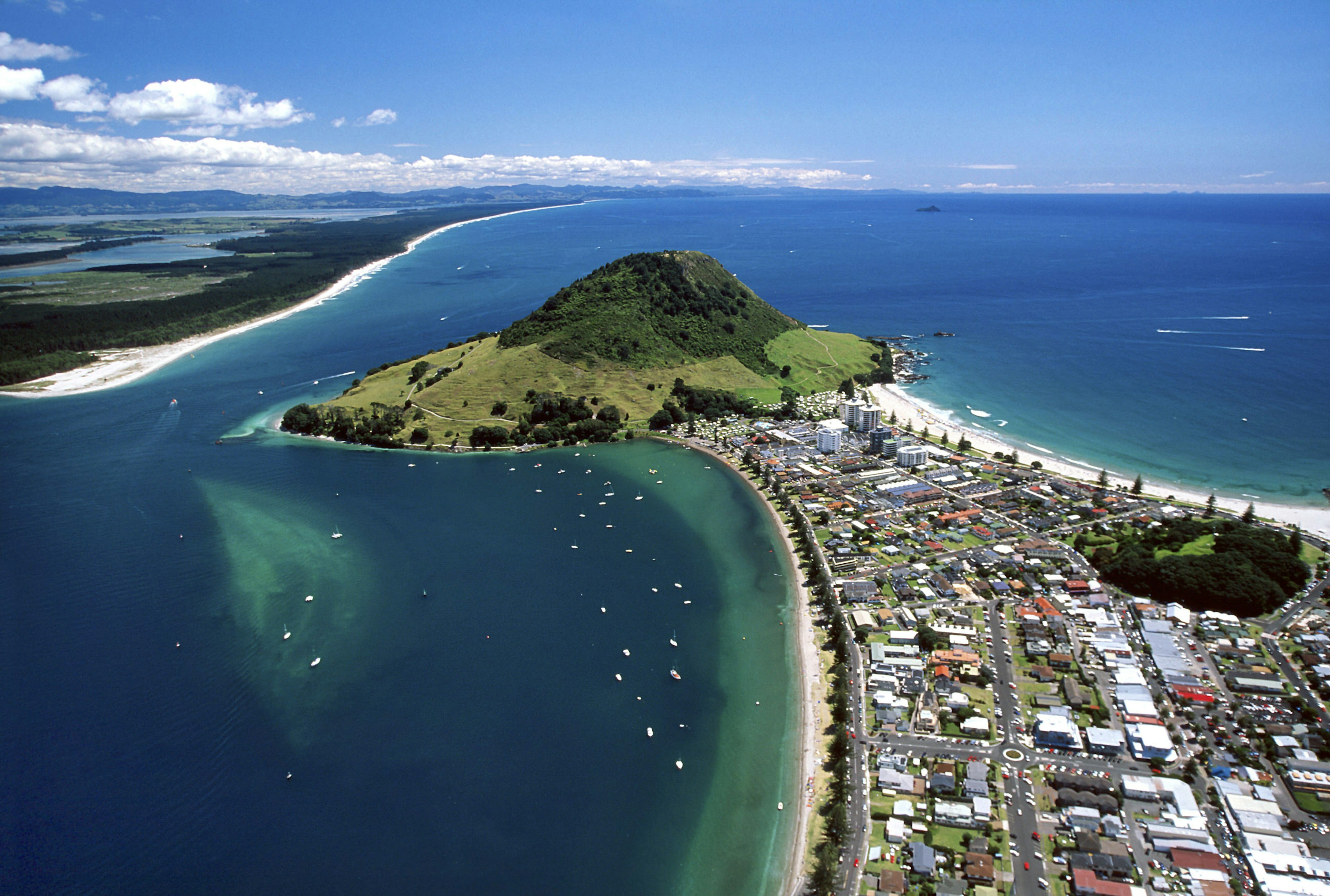 An aerial view of a coastline. There is a strip of land, covered in buildings and houses, with white, sandy beaches lining either side. At the land's end, there is a small mountain, covered in grass and lush green trees. All around is azure blue sea, dotted with boats.