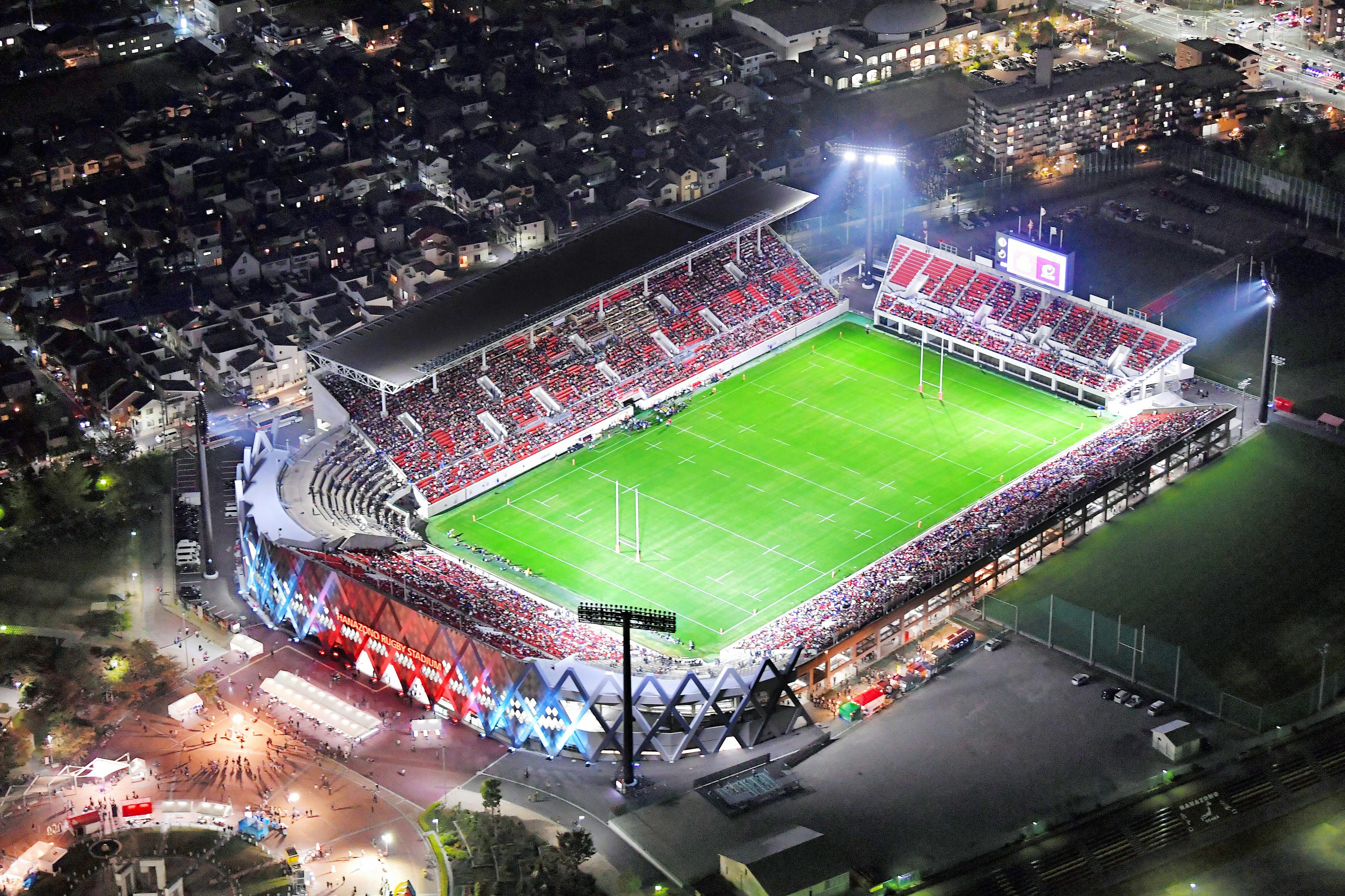 An aerial view of Hanazono Rugby Stadium at night with the pitch lit by floodlights