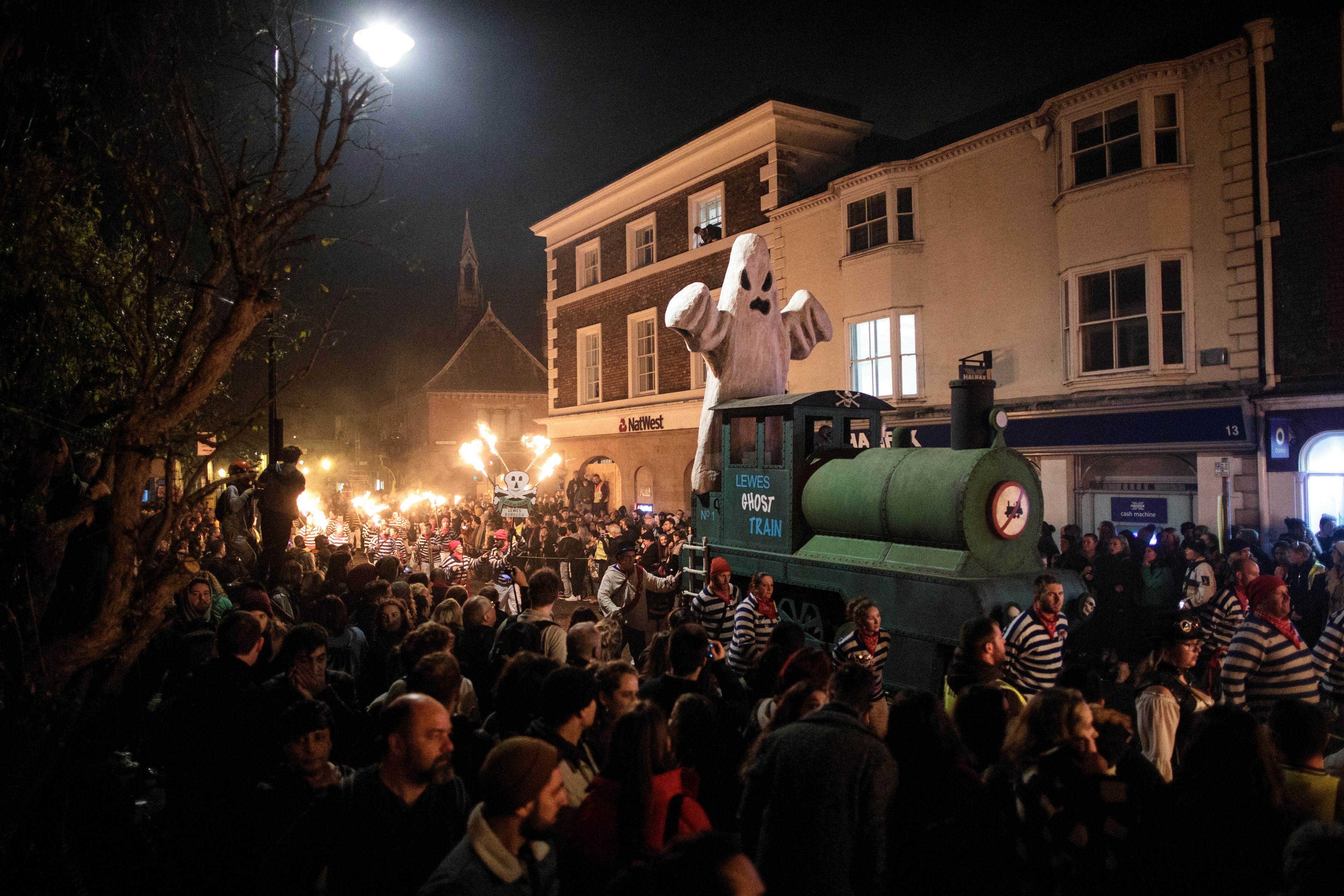 A tableaux featuring a green train with a ghost on it is led through a narrow street packed with revellers.