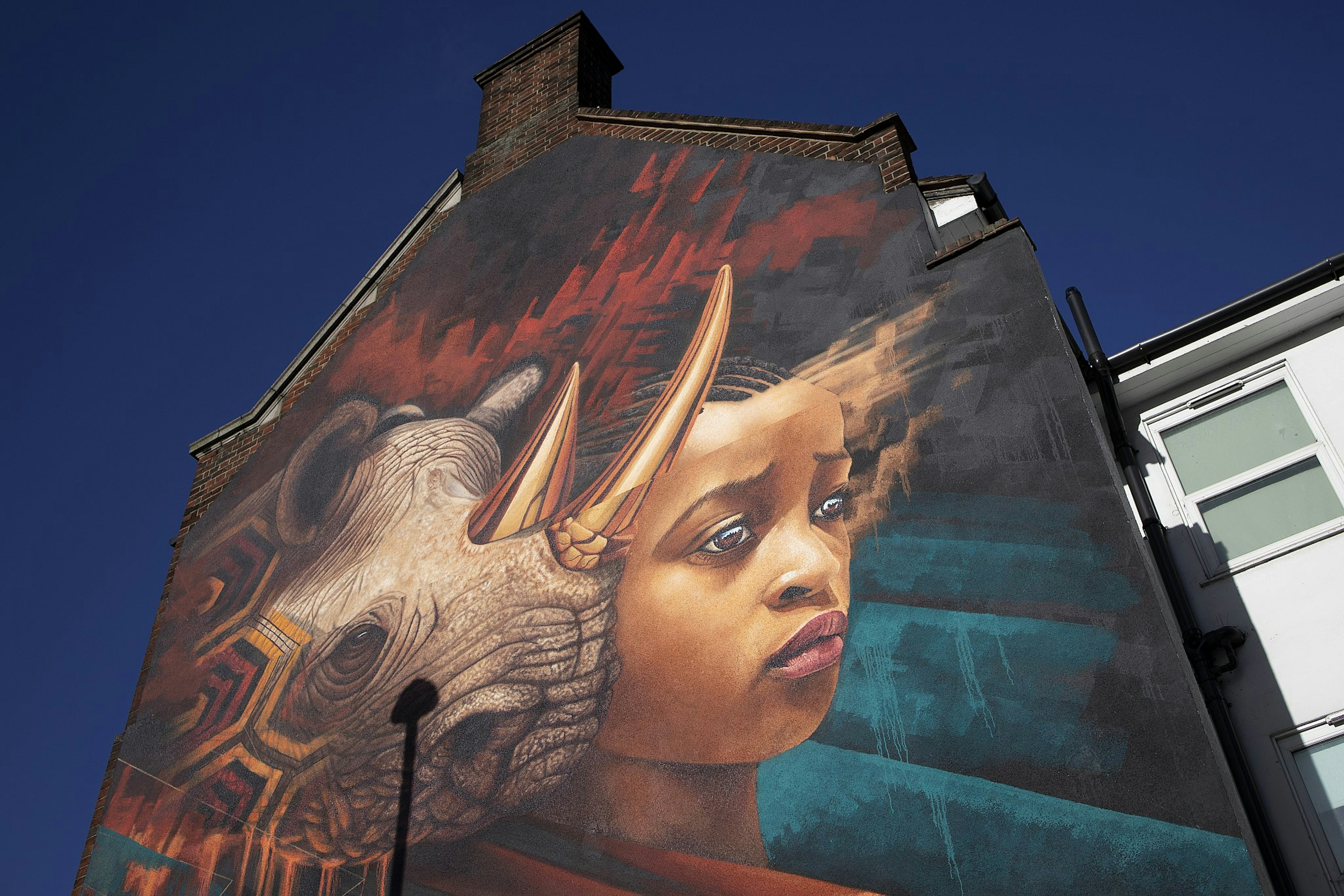 Looking up at the side of a house covered in a huge mural of a rhino next to a woman in front of an apocalyptic-looking scene with an angry red sky.