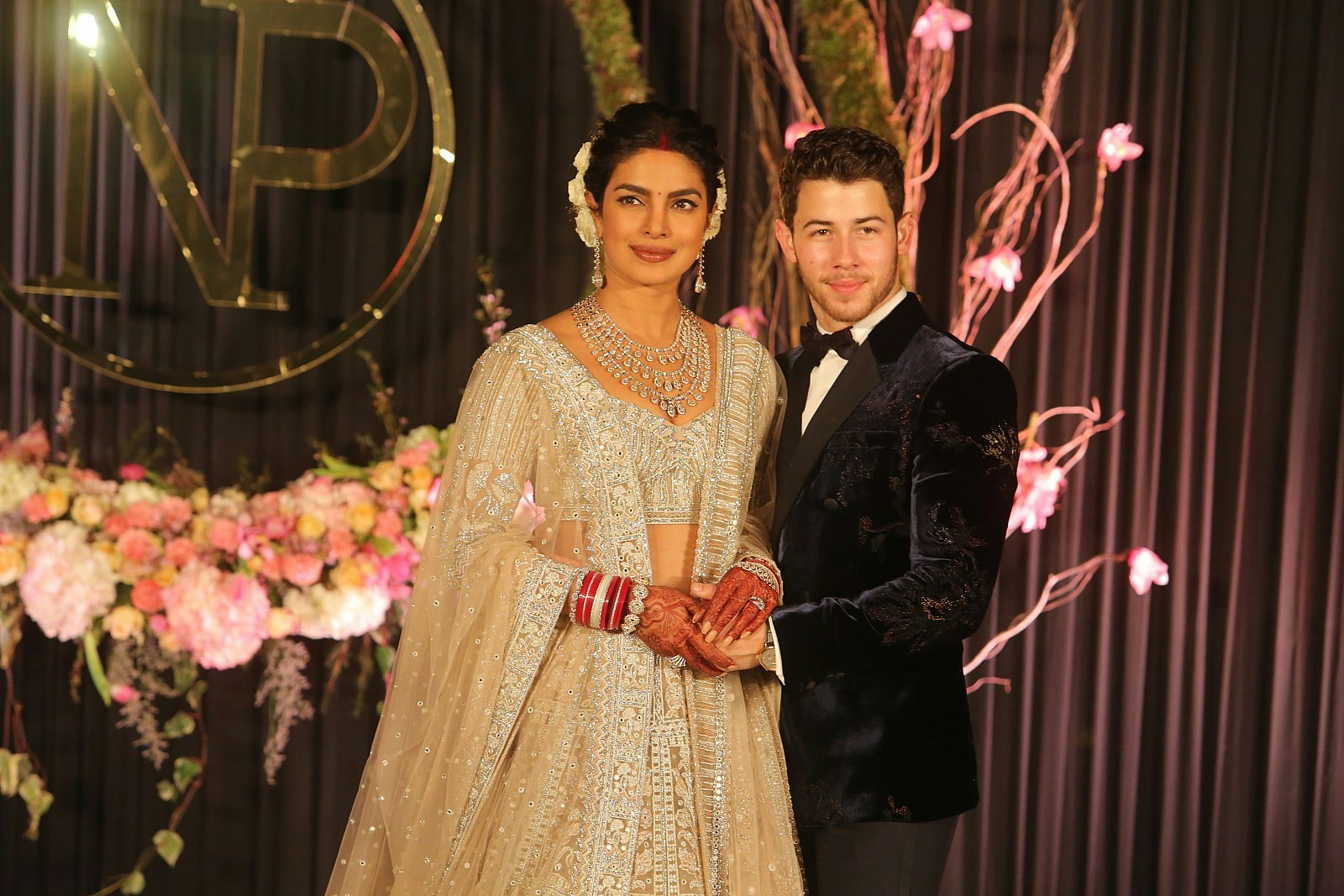 Priyanka Chopra and Nick Jonas at their wedding reception; she is wearing an elaborately beaded sari and necklace and he is wearing a dark velvet tuxedo.