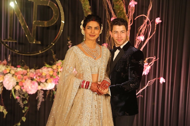 Priyanka Chopra and Nick Jonas at their wedding reception; she is wearing an elaborately beaded sari and necklace and he is wearing a dark velvet tuxedo.