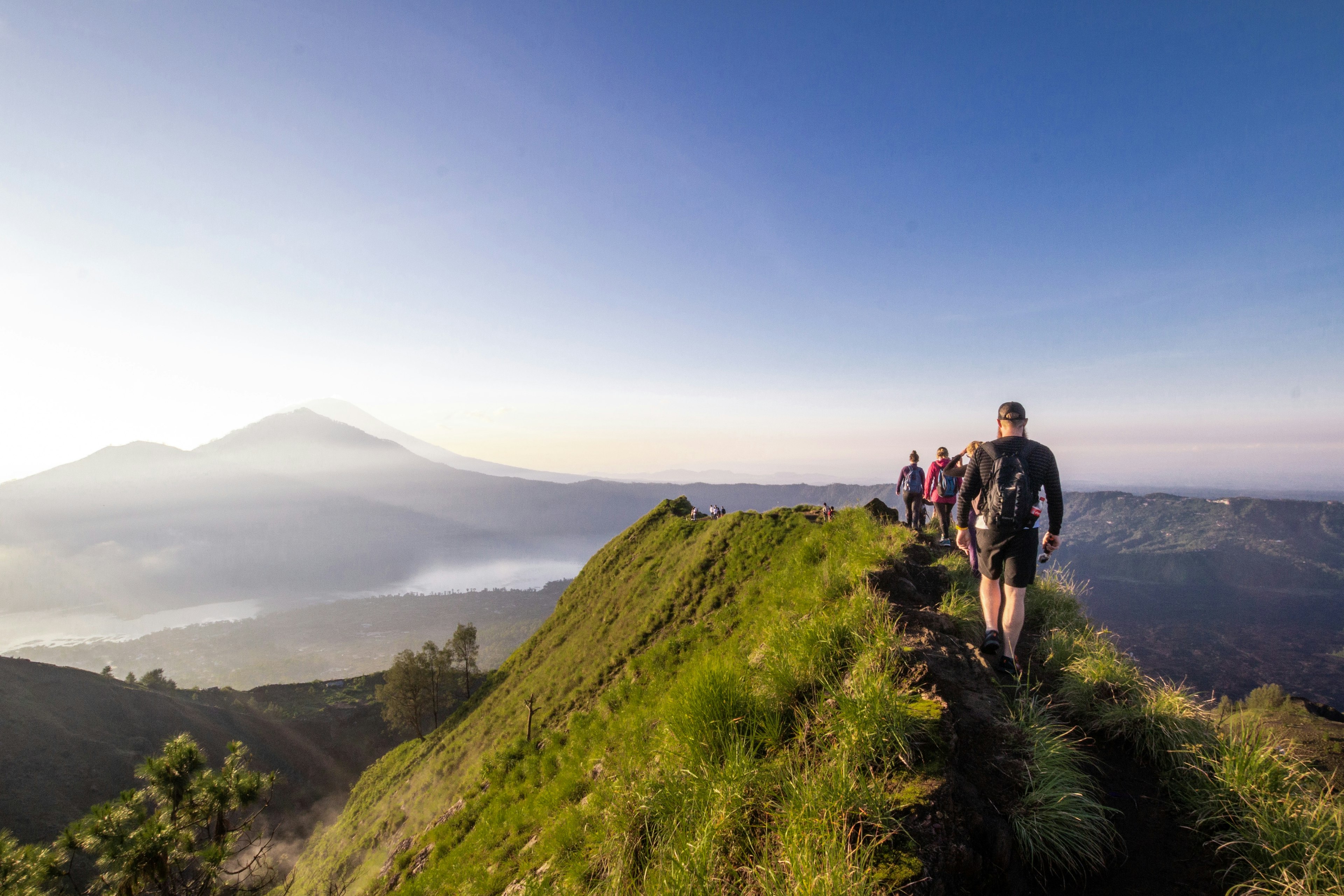 A group of hikers follow a narrow trail atop a mountain ridge in single file. Other peaks, covered in mist, are visible in the distance