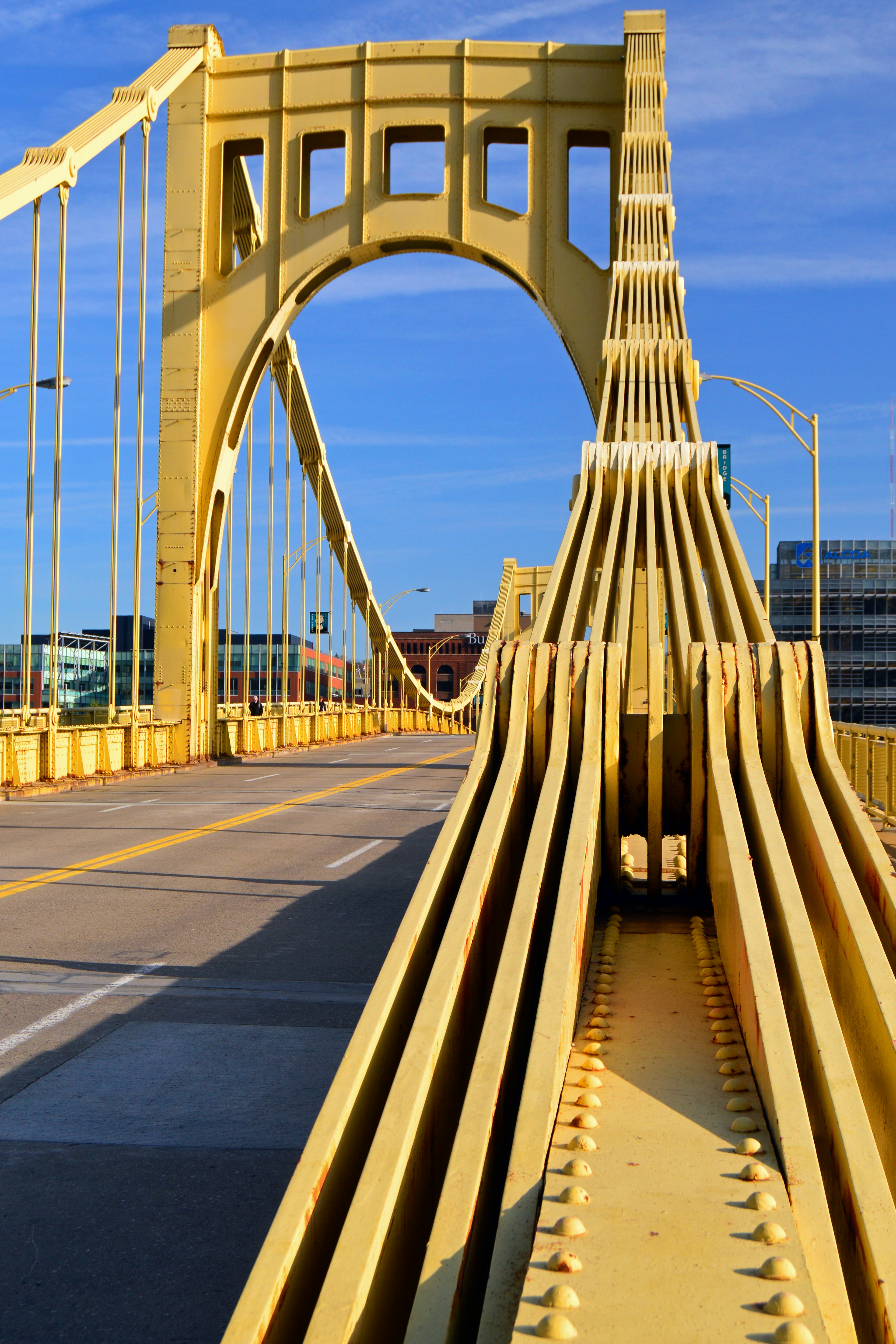 A close-up of one of the main arches of a huge yellow steel bridge. There are no vehibles on the four-lane road which crosses it.