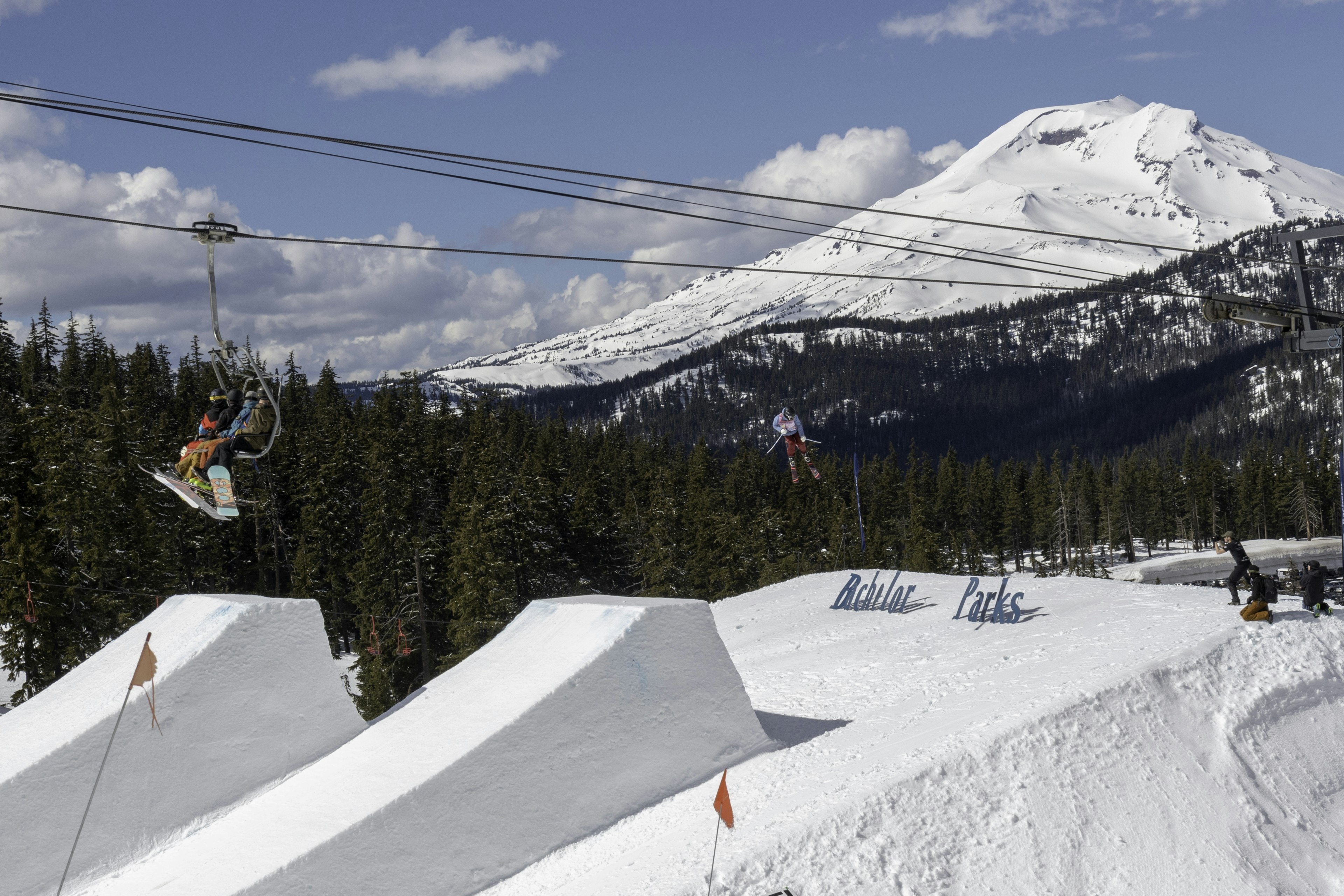 A skier is poised in mid air after taking a jump off one of several massive snow ramps at Mt. Bachelor, as snow boarders in colorful snow suits cruise by on a ski lift.