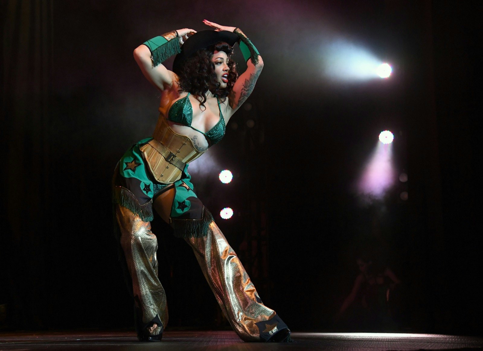 A woman in a green top and chaps performs burlesque in a spotlight