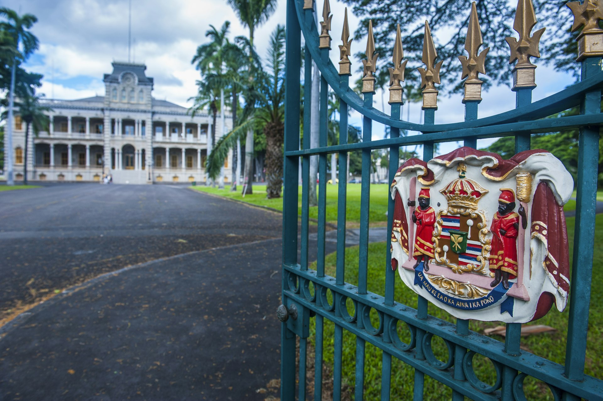 A crest on an exterior gate that leads up to a grand palace surrounded by palm trees