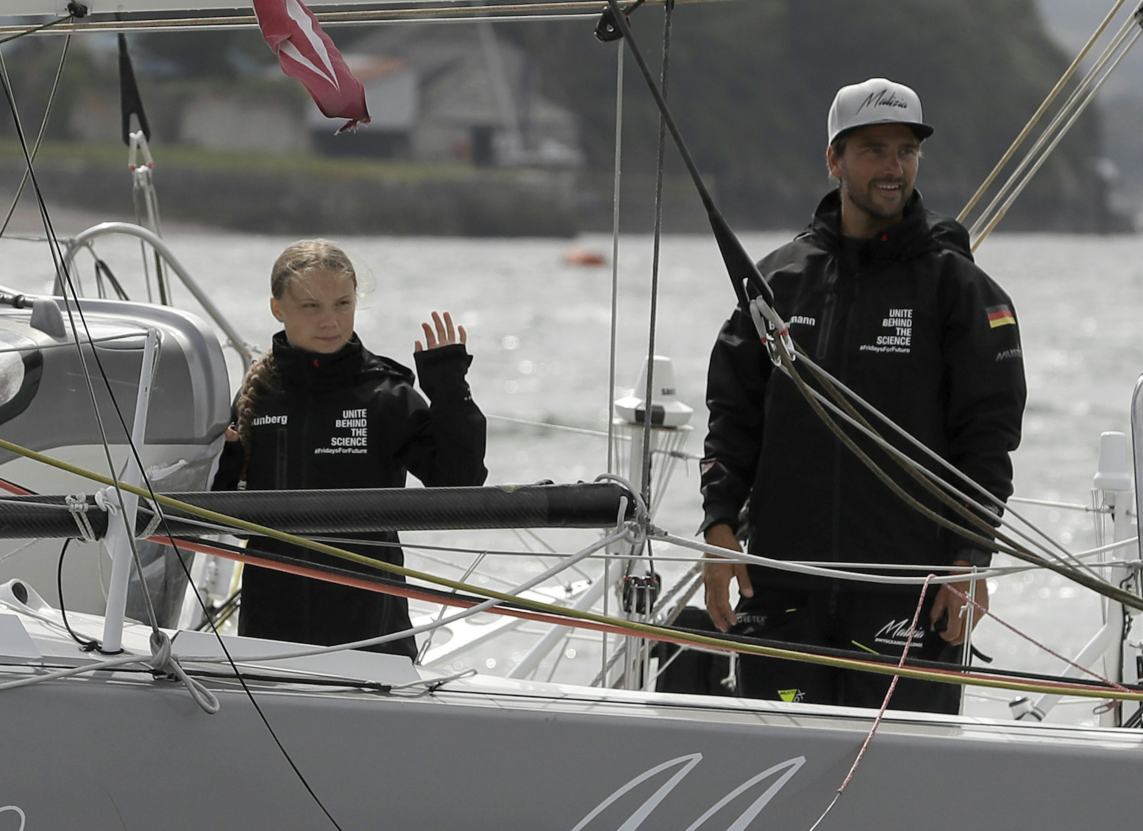 Climate activist Greta Thunberg stands on a solar powered sailboat ahead of her voyage to the 2019 United Nations Climate Action Summit. She wears a black windbreaker with the words United Behind the Science printed in white. She stands next to a man wearing a similar jacket, with the German flag on the left sleeve and a white ball cap on his head.