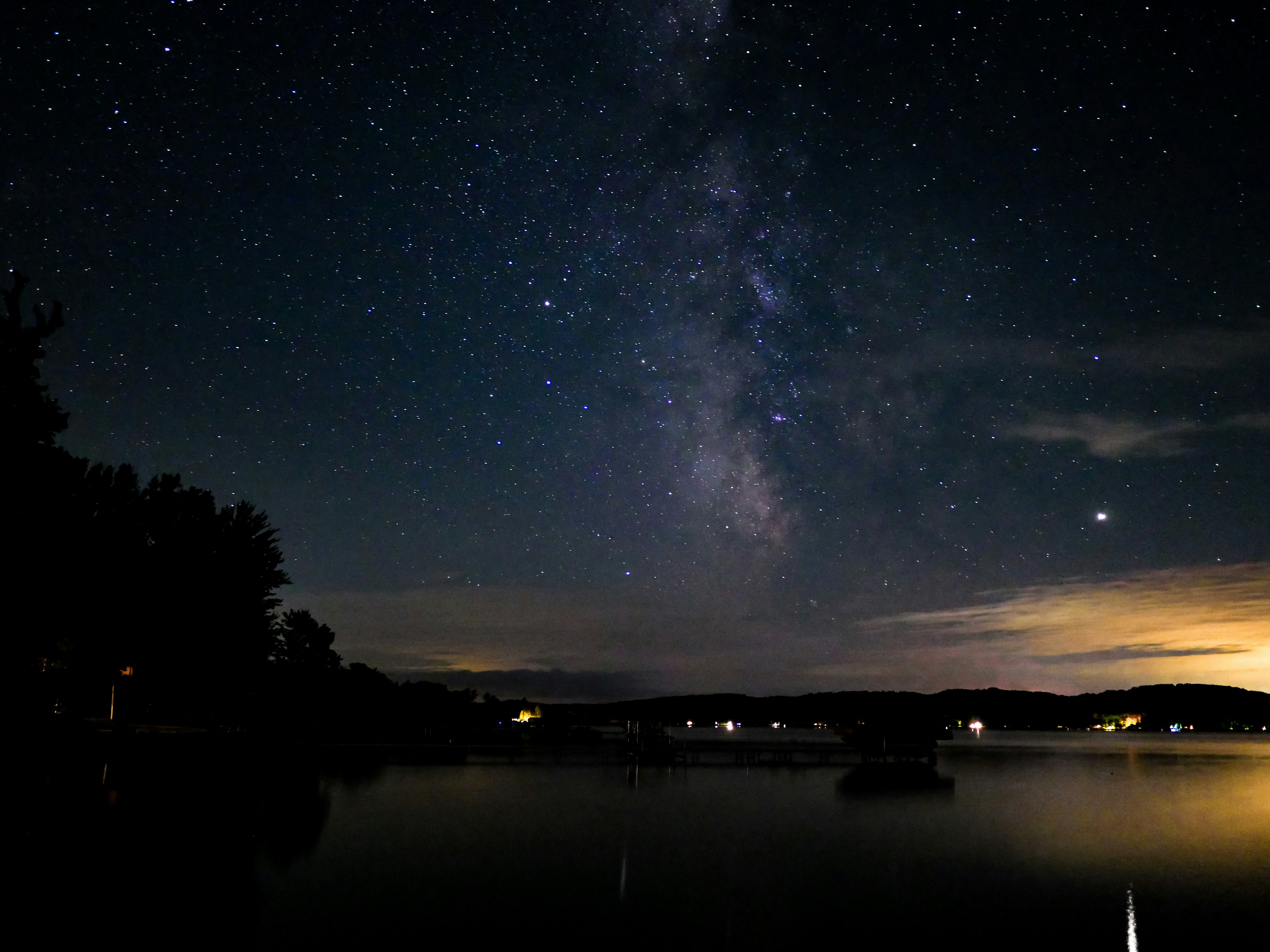 A long exposure photo shows the stars and Milky Way glittering against a dark blue sky over a Michigan lake, with warm amber lights from a few houses and minimal development lower down in the frame hi-lighting the shoreline.