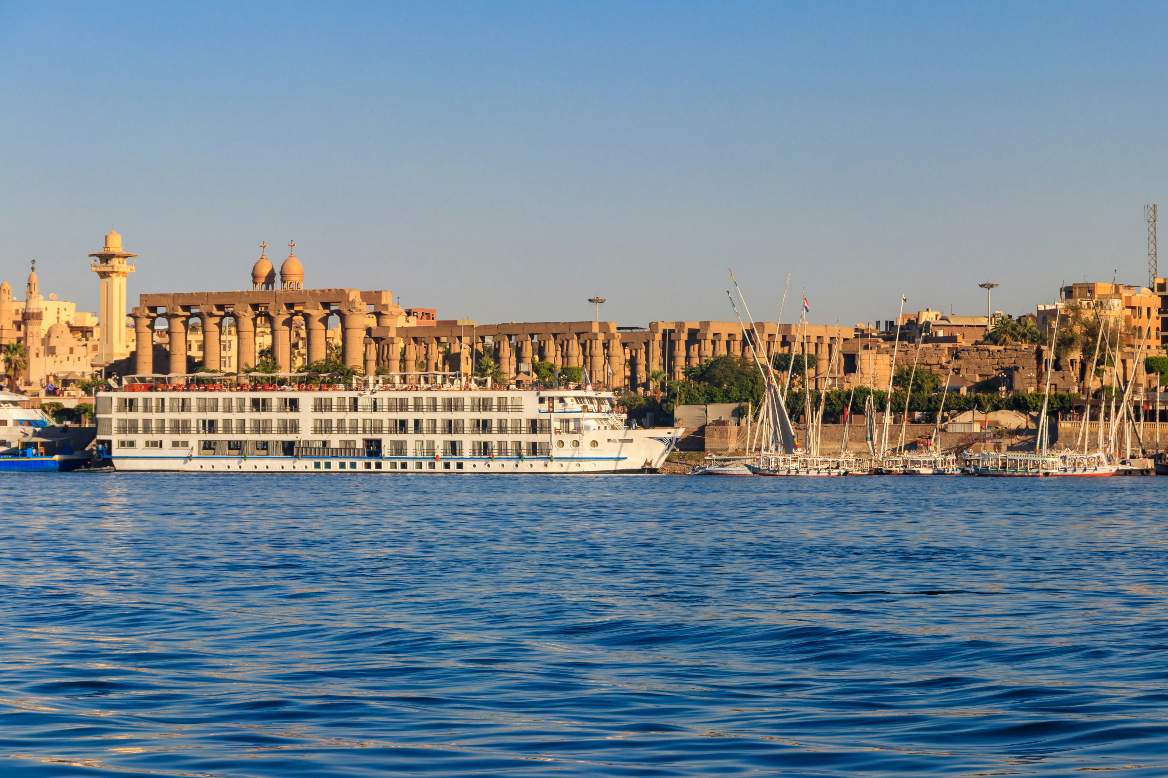 A large cruise boat and many small sailboats are moored at the side of a river in front of a large ancient temple complex