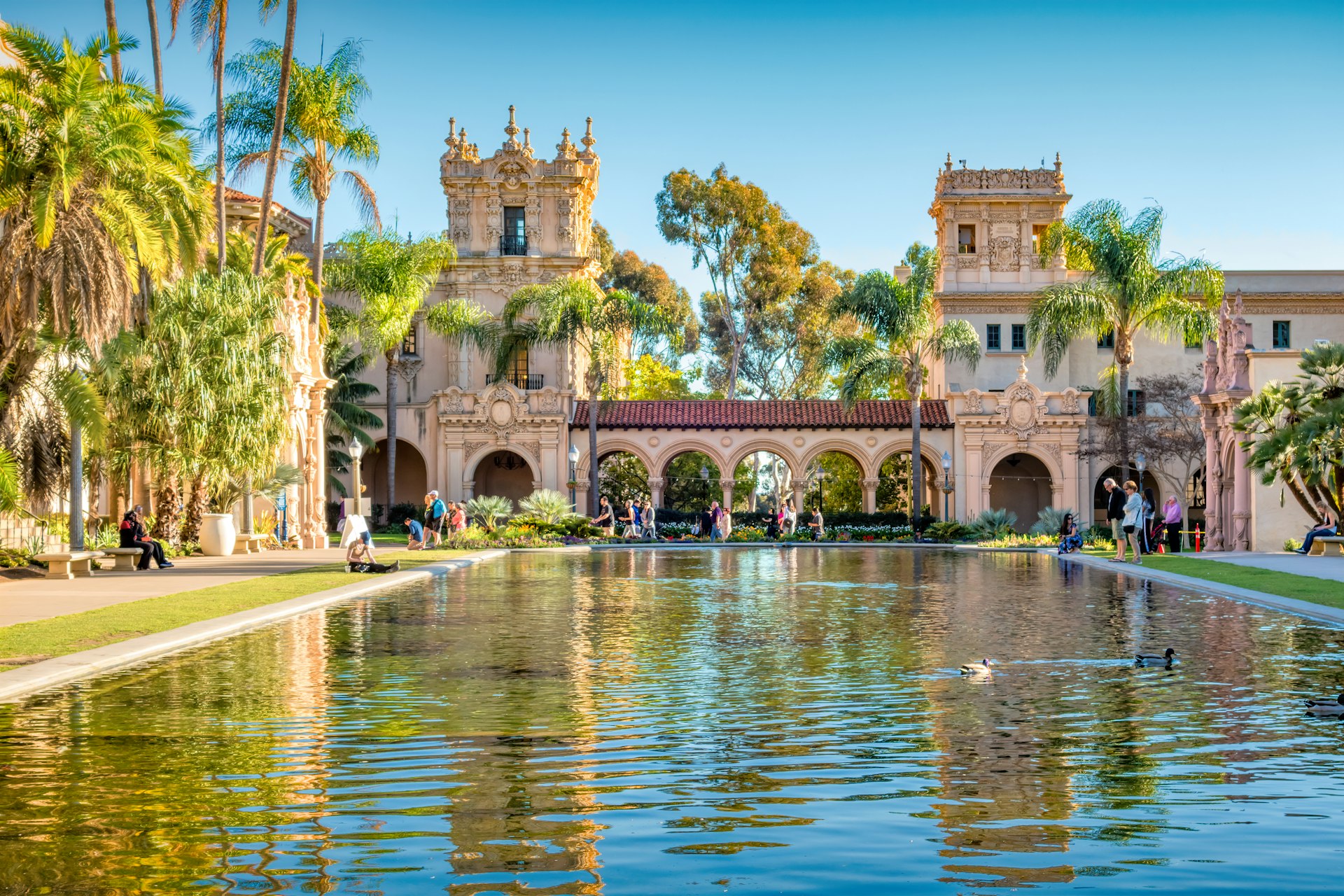 People walk around a large rectangular ornamental pond in front of a building with two towers