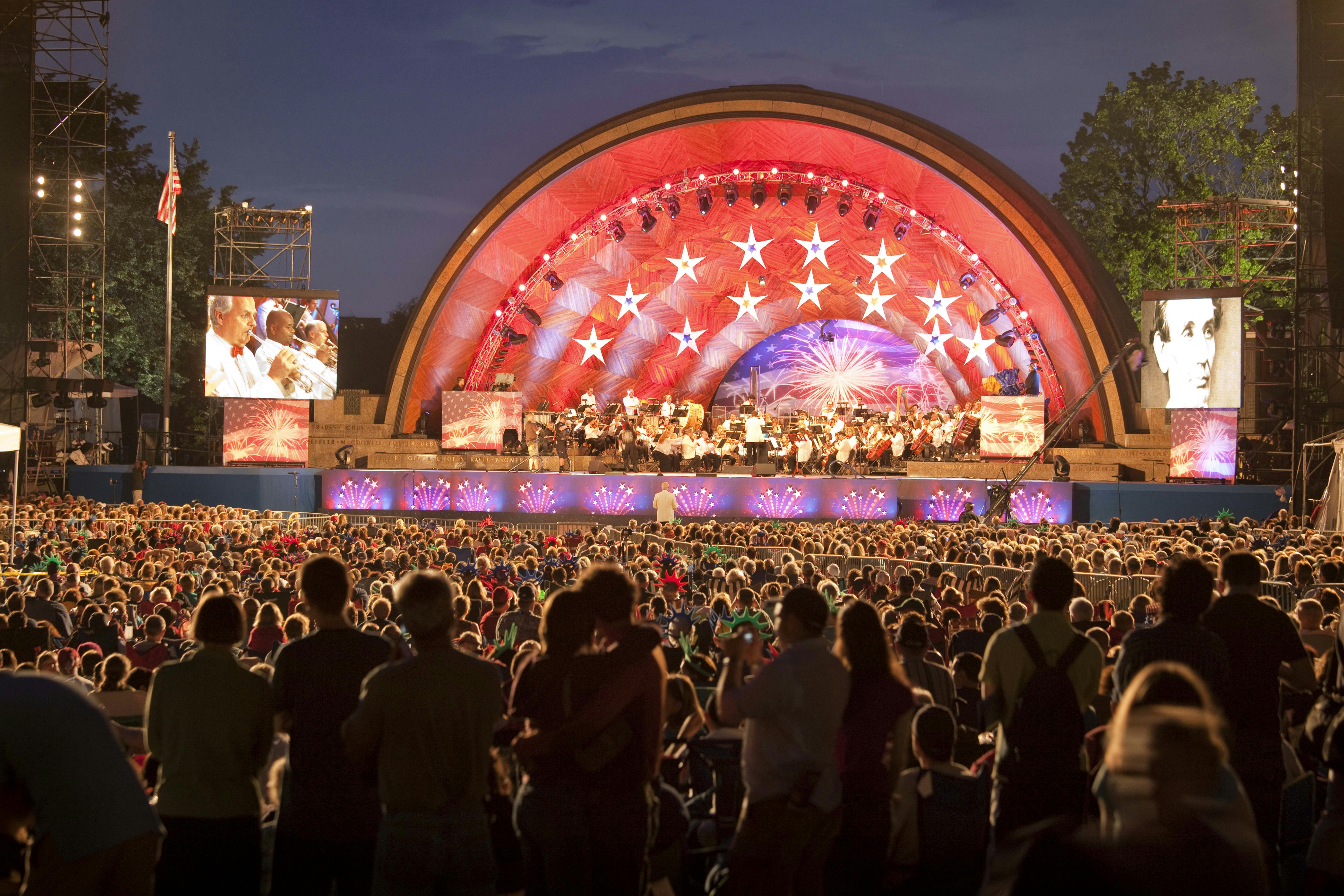 A crowd of people watch the brightly colored stage as the Boston Pops perform at the Hatch Memorial Shell in Boston