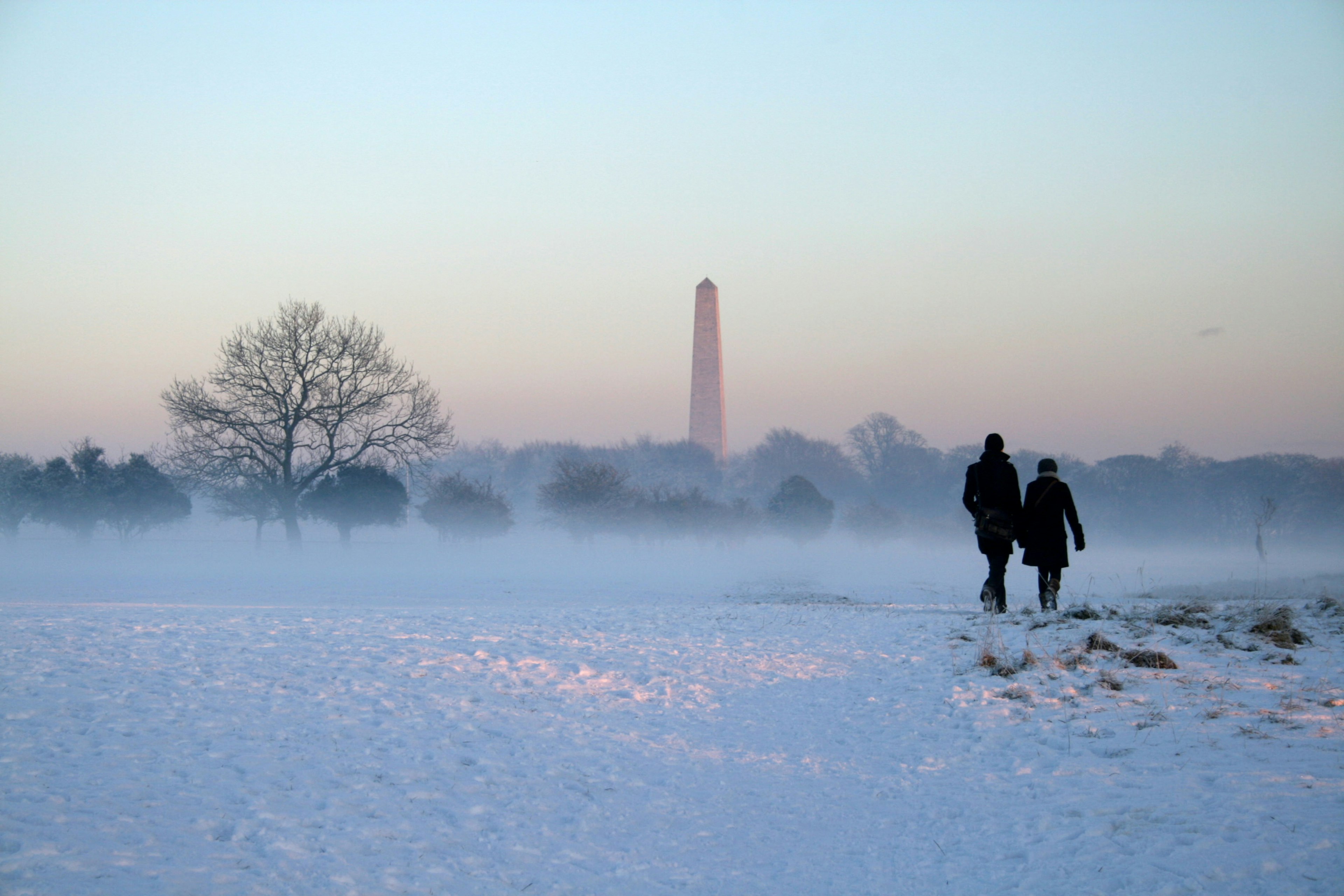 Phoienix Park Dublin in winter. Two people walk across the park which is covered in snow towards a pink horizon and a lone stone tower.