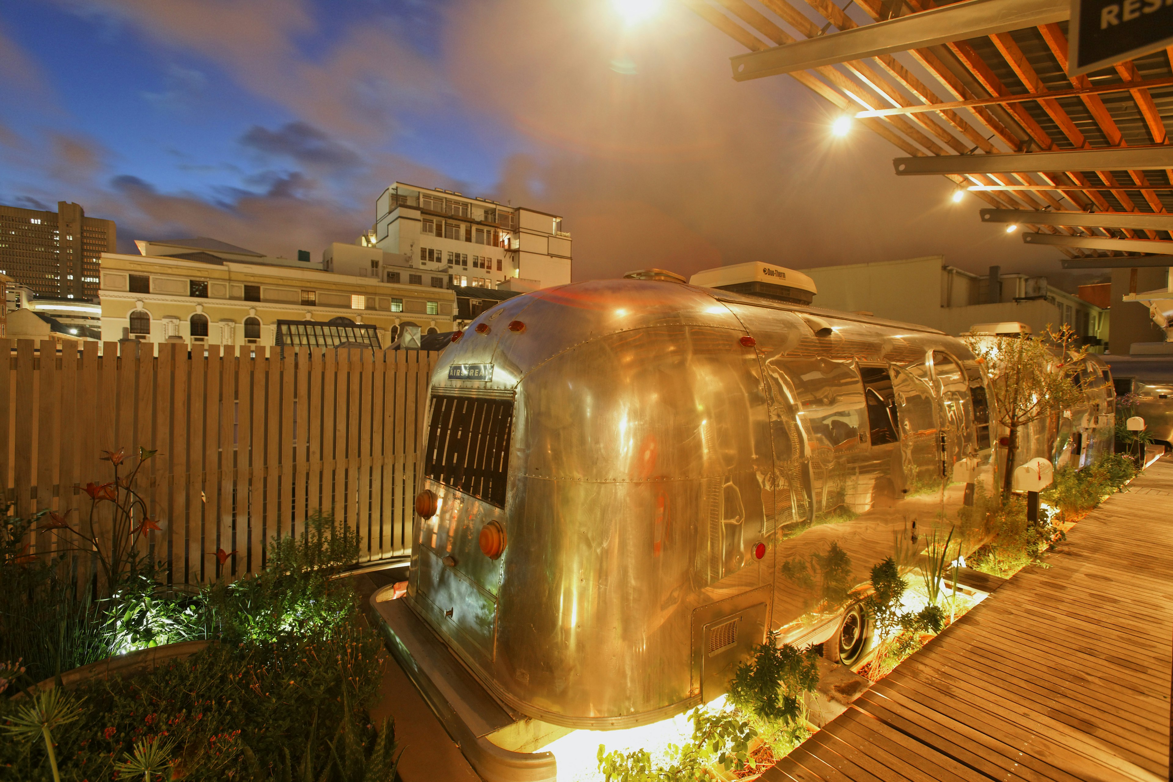 Gleaming silver Airstream trailers have a golden glow thanks to the warm wood fence, deck, and ceiling over the carefully manicured sites where they're parked at the Grand Daddy Hotel in Cape Town. Two trailers parked in a row have lights beaming up on them from the ground and shining down from the overhanging roof above. Behind them and the fence, white multi-story buildings hint at the surrounding neighborhood.