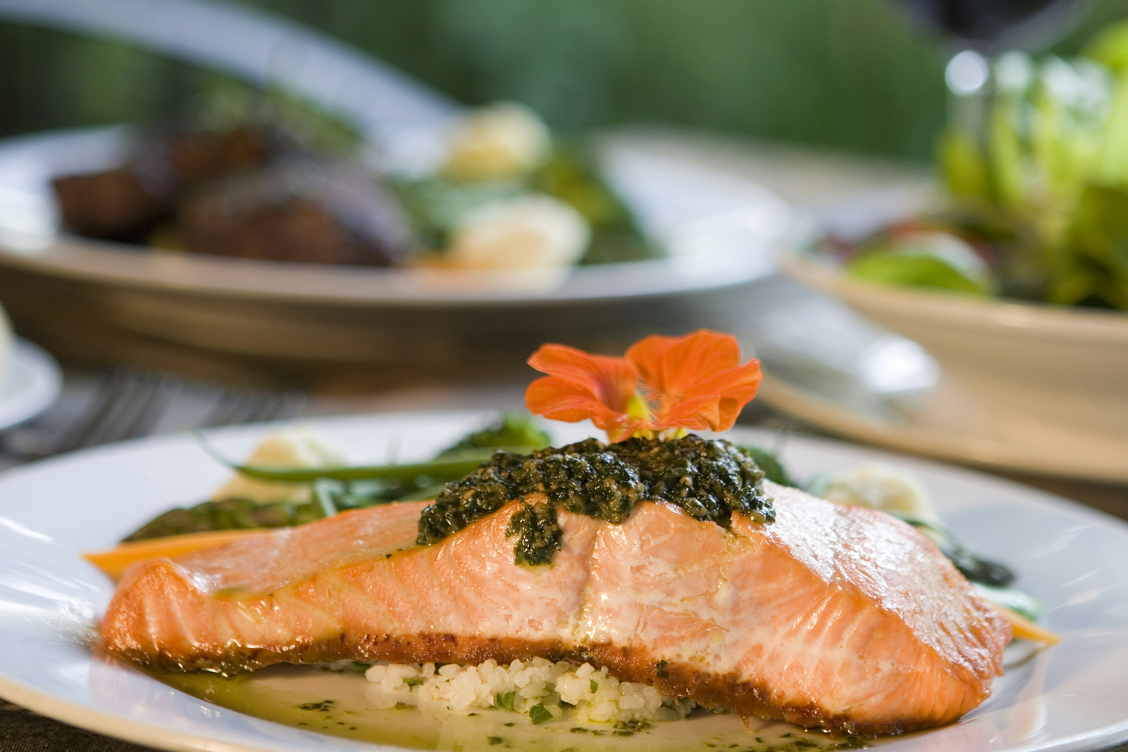 A fresh piece of orangey pink salmon is topped with a dark green herbed chimichurri and a bright orange flower over a bed of couscous and broth. Other plates heaped with vegetables sit in the background, forming blurred green tones.