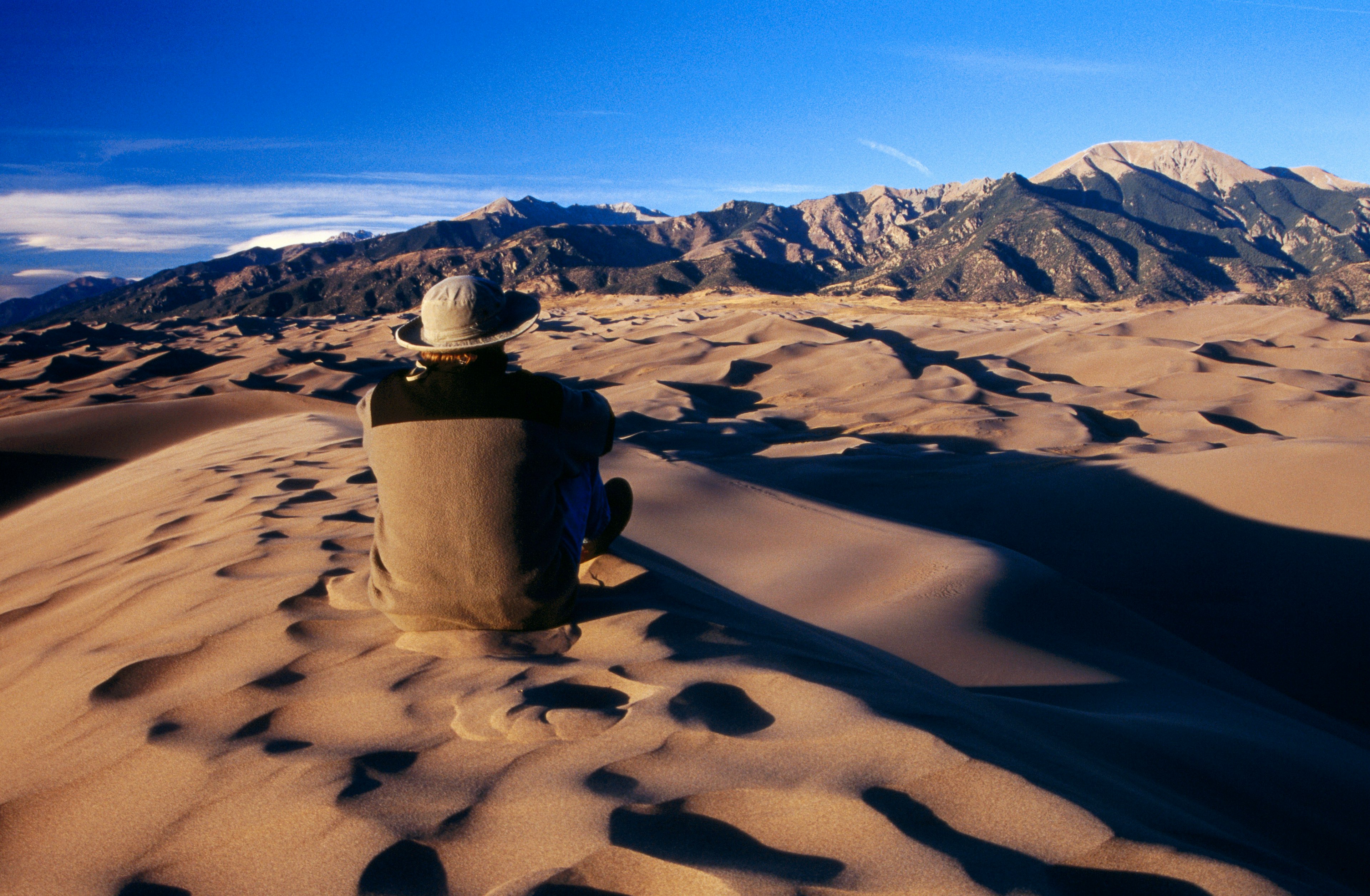 A man in a beige fleece jacket and a twill bucket hat sits with his back to the camera, blending into the sand dune on which he is perched, staring at the vast Sangre de Cristo Mountains in the distance.