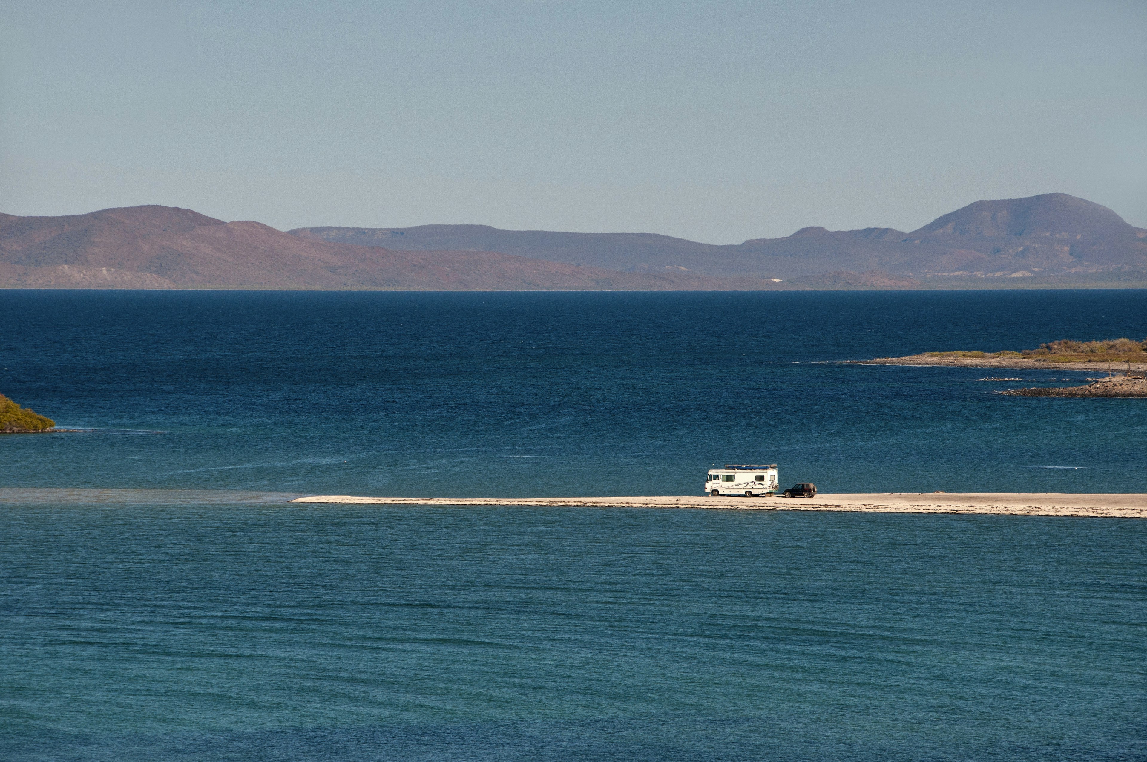 A vast blue sea surrounded by mountains is bisected by a spit of sand on which sits an RV