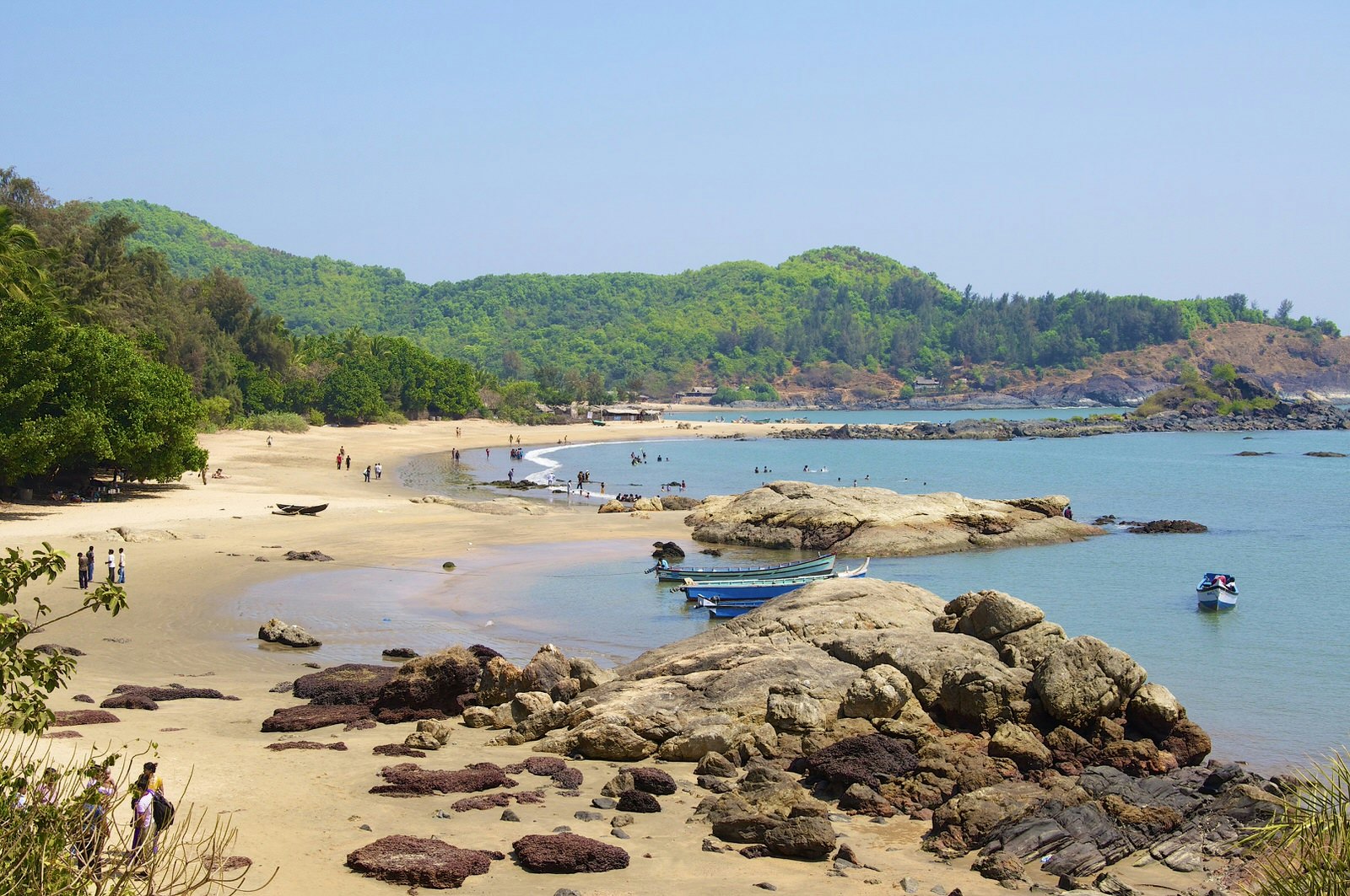 Late afternoon shot at Om beach in Karnataka, India. A few groups of people walk in the sand, which is backed by green forest, and several boats bobs in the shallow water