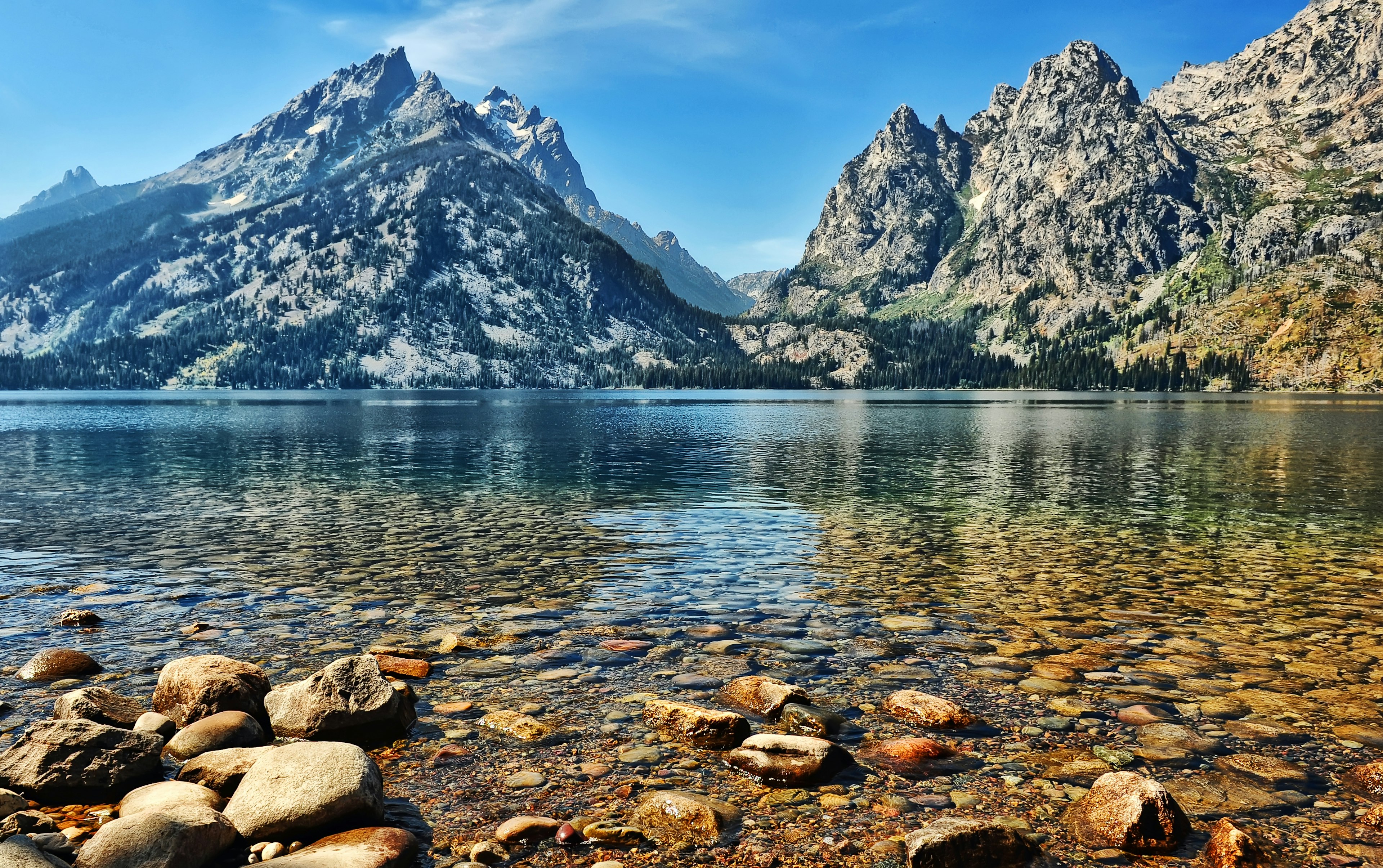 The ragged peaks of Grand Teton National Park rise steeply over Jenny Lake. The massif on the left is deep blue in the shadows and where trees cover its flanks, while the bright sunshine hi-lights its light grey rocky face. On the right, another peak is more brightly lit, appearing a light tan color with thin streaks of bright green vegetation. The lake itself is smooth and calm, a mixture of blues and greens, while in the extreme foreground the large, flat, stones on the shoreline are rust colored and grey