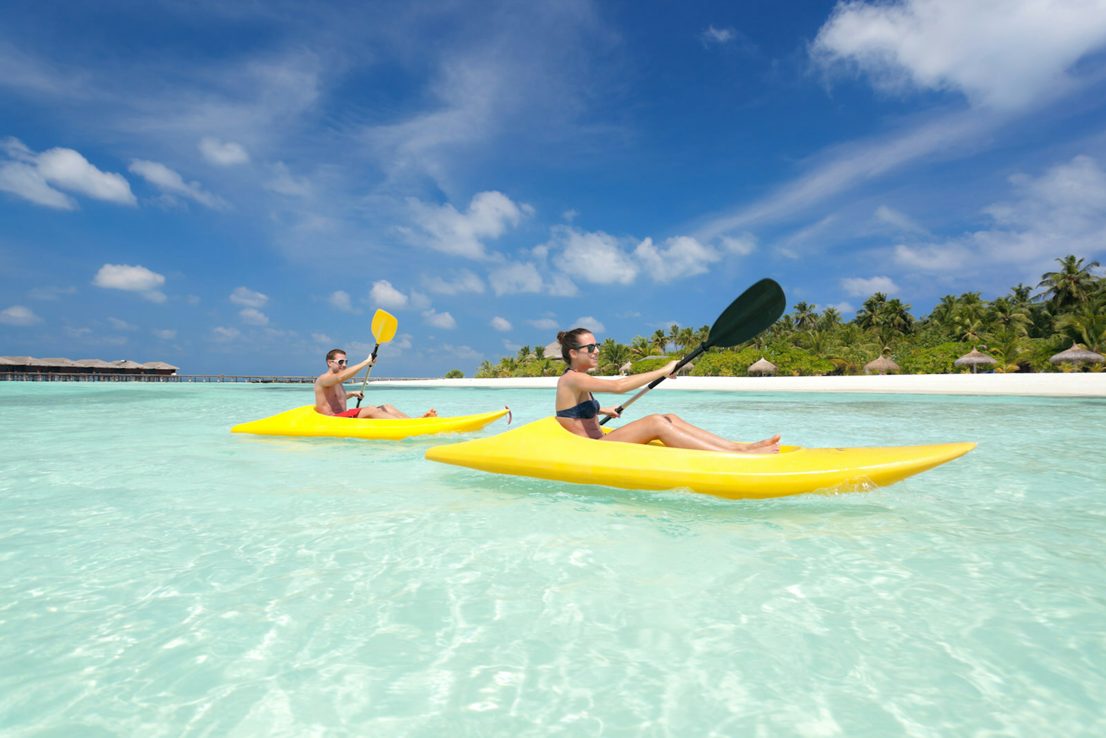 A couple in yellow kayaks paddle through crystal-clear water in the Maldives, with blue skies above them and a row of sunshades in the white sand in the distance.