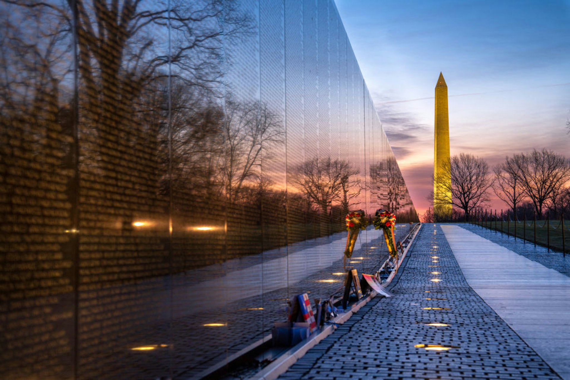 Sunrise is reflected in "The Wall", a large memorial listing names of those lost in the Vietnam War. A large stone obelist-shaped monument is in the background