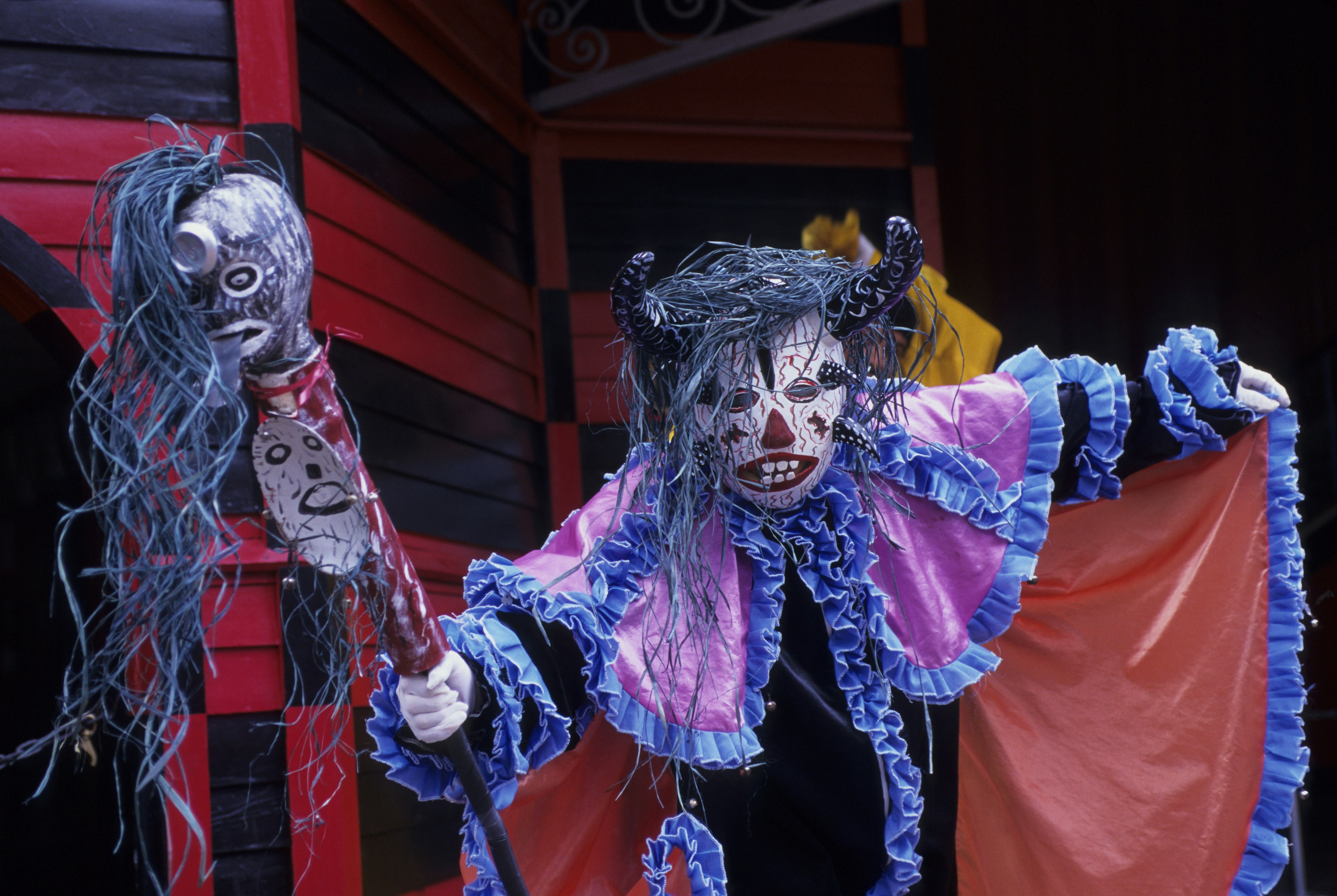 A performer wearing a red and blue cap, wearing a horned face mask and carrying a staff leans menacingly towards the viewer, part of Afro-Puerto Rican festival custom