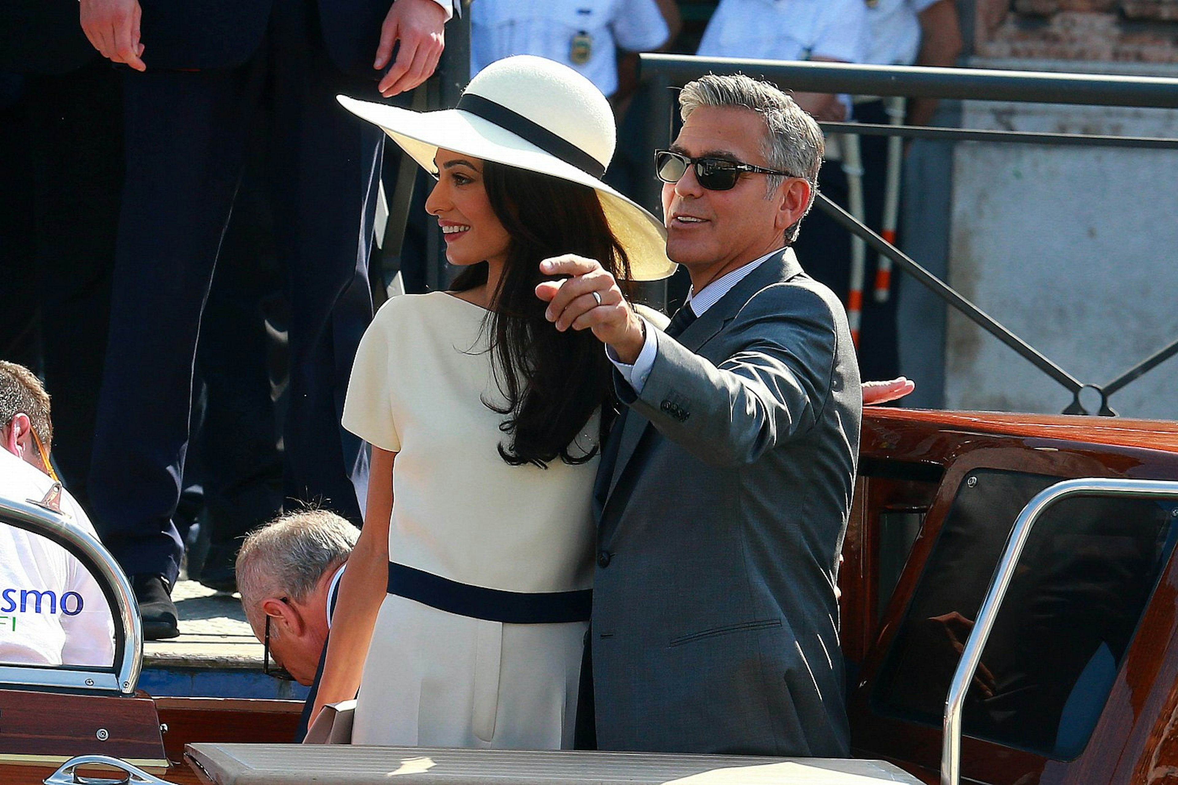 George and Amal Clooney arriving at their civil ceremony in Venice on a boat; she is wearing a cream trouser suit and wide-brimmed cream hat and he is wearing a dark grey suit.