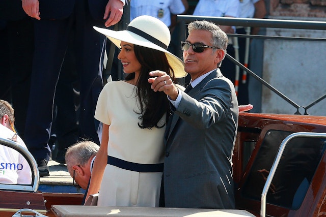 George and Amal Clooney arriving at their civil ceremony in Venice on a boat; she is wearing a cream trouser suit and wide-brimmed cream hat and he is wearing a dark grey suit.
